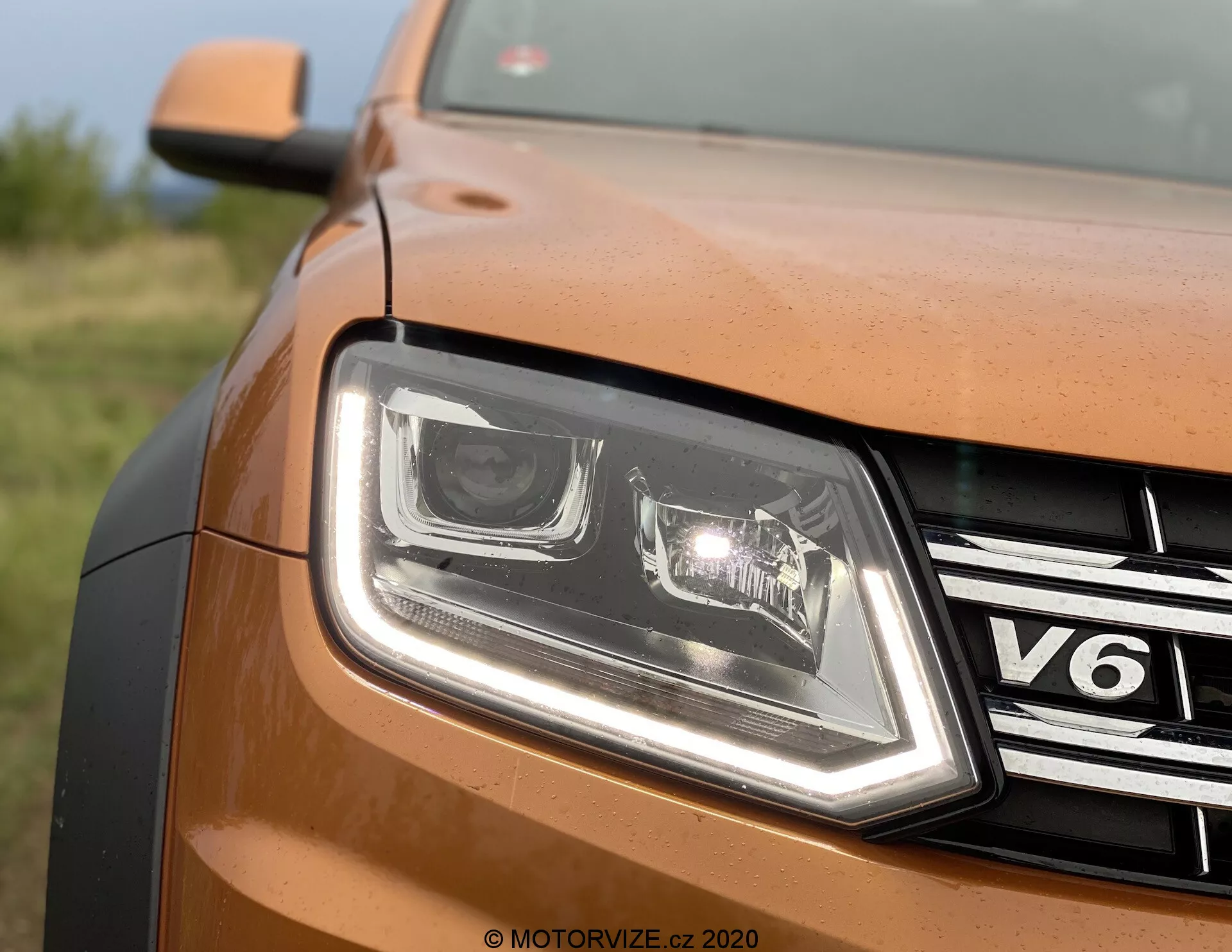 Detailný záber čiastočného predného pohľadu na bronzové alebo medené auto VW Nutzfahrzeuge Amarok 2016 z ľavej strany. Obrázok ukazuje moderný dizajn LED svetiel s charakteristickými dennými svetlami, chrómovanú mriežku s odznakom 'V6', kovový lak karosérie, obrysovanú hranu kapoty a vodoznak 'MOTORVIZE.CZ 2020', čo naznačuje pôvod z českého recenzného webu.