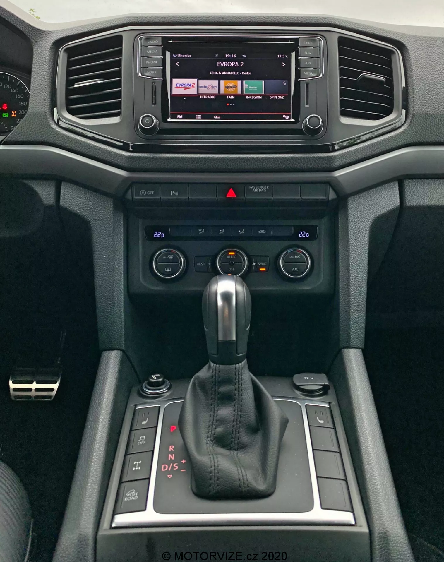 Detailný pohľad na modernú stredovú konzolu VW Amarok 2016, zameraný na infotainment systém a radiacu páku. Infotainment displej naznačuje dotykové ovládanie, pravdepodobne zobrazuje rádio alebo mediálny výstup. Nižšie sú horizontálne vetracie otvory obklopené mäkkými materiálmi s textúrovaným dizajnom. Pod vetrákmi sú ovládacie prvky klimatizácie a prepínače na nastavenie teploty a prúdenie vzduchu. Dominuje automatická radiaca páka v režime 'P', čo naznačuje automatickú prevodovku s možnosťami ručného radenia. Vedľa radiacej páky sú malé úložné priehradky a otočný ovládač pravdepodobne pre infotainment alebo systém jazdných režimov. Dizajn interiéru zdôrazňuje funkčnosť a pohodlie.