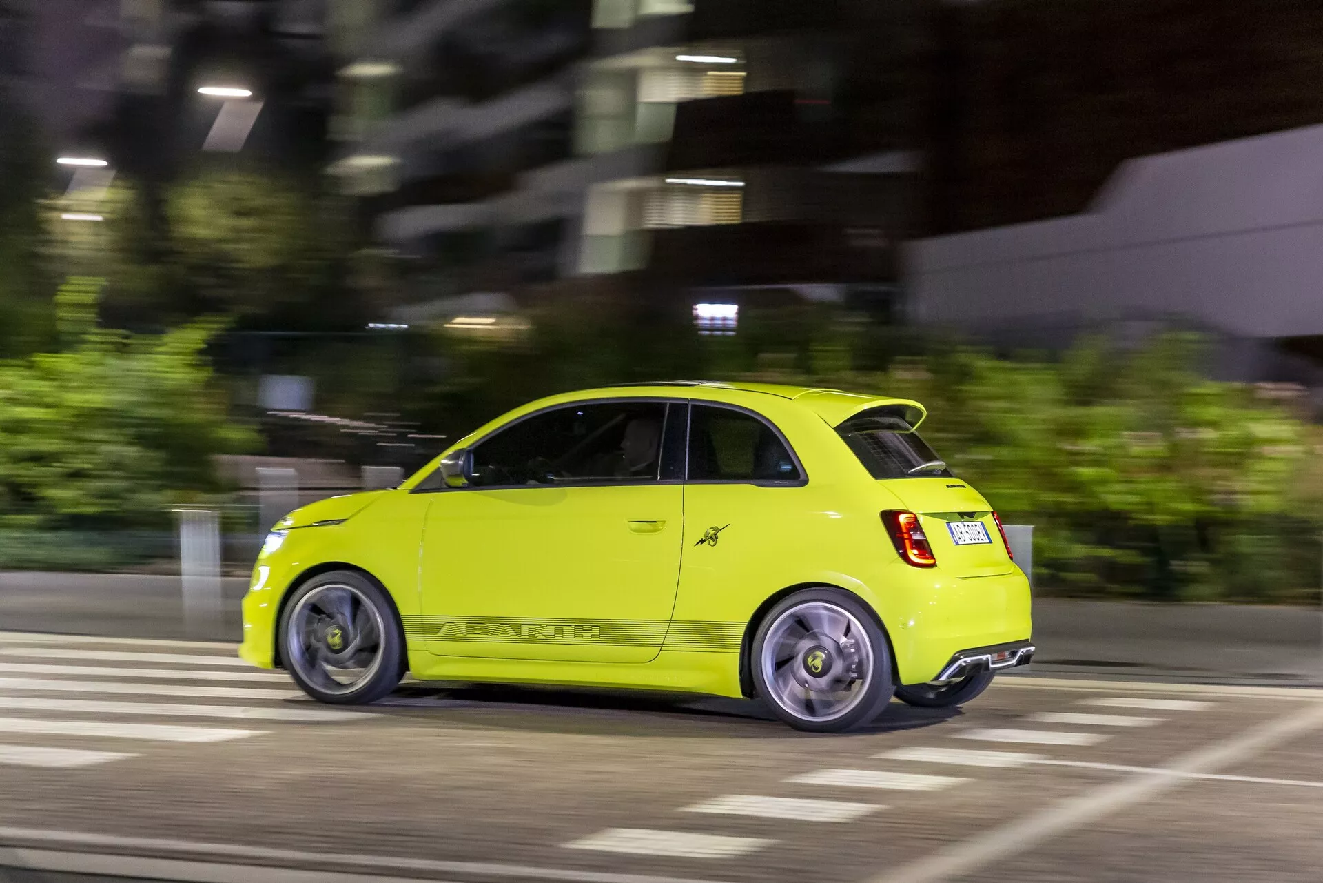 Dynamická nočná fotografia žiarivo žltého auta Abarth 500-595-695 2023 druhej generácie z bočného zadného pohľadu, zachytená technikou sledovania pohybu. Kľúčové vlastnosti zahŕňajú kompaktný hatchback dizajn, rozsvietené zadné svetlá, rozmazané zliatinové kolesá pohybom, lesklú žltú farbu, zadný emblém a mestské prostredie so zebrovým priechodom, ktoré zvýrazňujú jeho dizajn, bezpečnosť, výkon a aerodynamiku pre mestskú mobilitu.