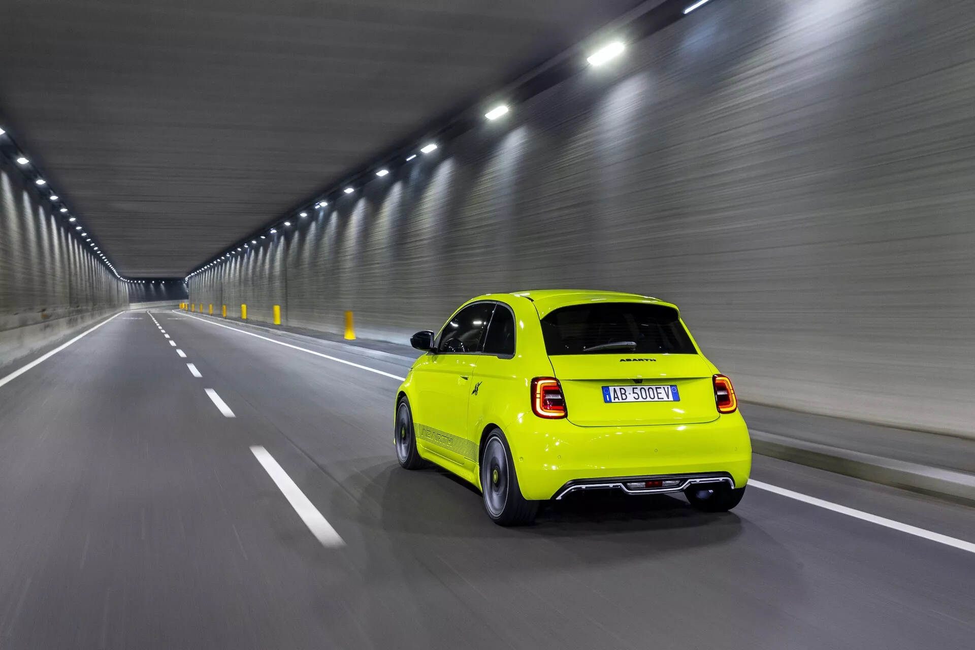 Dynamický zadný pohľad na jasne žlté vozidlo Abarth 500-595-695 2023 (2. generácia) jazdiace cez tunel. Nachádza sa v pravom pruhu, mierne zarámované doľava s viditeľným okolím tunela. Fotografované z nasledujúceho vozidla, uhol kamery je nízky, ukazuje zadnú a bočnú časť vozidla, dizajn zadných svetiel, zadné stierače, výfukový systém a zadný difuzér. Kolesá sú mierne rozmazané, čo naznačuje pohyb. Vnútorné osvetlenie tunela rovnomerne osvetľuje auto, zvýrazňuje jeho kompaktný dizajn a aerodynamické prvky.