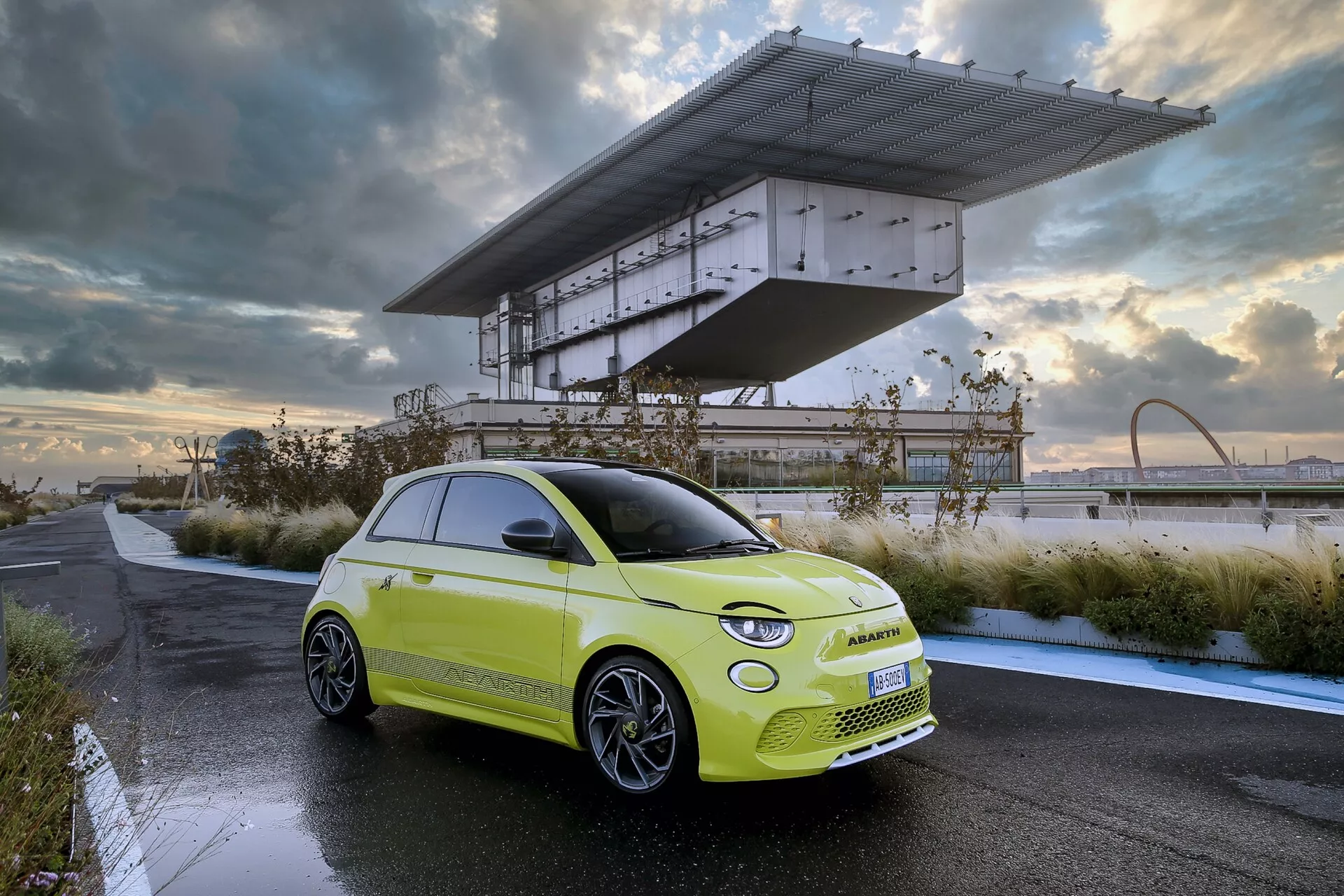 Moderné hatchback auto v trojštvrťovom prednom pohľade, zobrazené vonku na spevnenej ploche. Má jasne limetkovú zelenú karosériu s kontrastnou čiernou strechou, zvýrazňujúcou prednú masku, LED svetlomety, aerodynamické línie kapoty, viaclúčové kolesá a elegantnú bočnú siluetu. Súčasný dizajn vozidla a prirodzené osvetlenie naznačujú svitanie alebo súmrak, s dynamickými mrakmi a vzdialenými architektonickými prvkami v pozadí. Obrázok zdôrazňuje pokročilé automobilové technológie a ekologický dizajn.