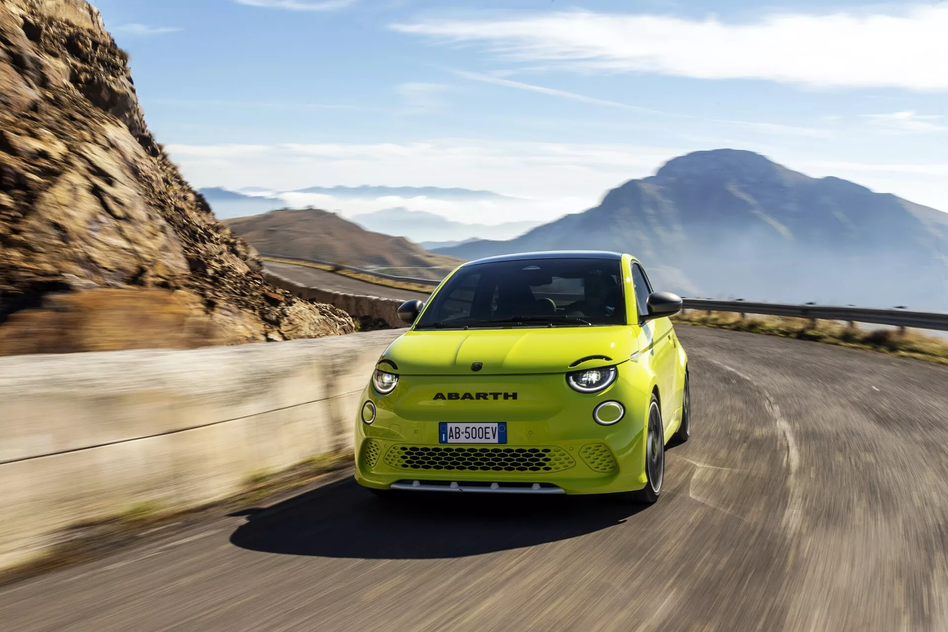 Obrázok zachytáva žlté auto Abarth 500-595-695 2023 (2. generácia) v pohybe, fotografované z nízkeho predného trojštvrťového uhlu. Tento uhol poskytuje detailný pohľad na dizajnové línie auta, vrátane jeho zaoblených svetlometov, charakteristickej prednej mriežky so stredovým odznakom, športových kontúr na kapote a aerodynamických línií po bokoch. Sú zreteľne viditeľné hliníkové kolesá a nízka pozícia kamery zdôrazňuje postoj a svetlú výšku vozidla. Auto je umiestnené na pravej strane obrázka, pohybujúce sa po zákrutej ceste, čo naznačuje dynamický pohyb vpred, s rozmazaným pozadím vytvárajúcim pocit rýchlosti. Scenéria zahŕňajúca hory a čistú oblohu zvýrazňuje dobrodružný a výkonnostný charakter auta, typický pre značku Abarth. ŠPZ je rozmazaná, zachováva súkromie a koncentruje pozornosť na vozidlo.