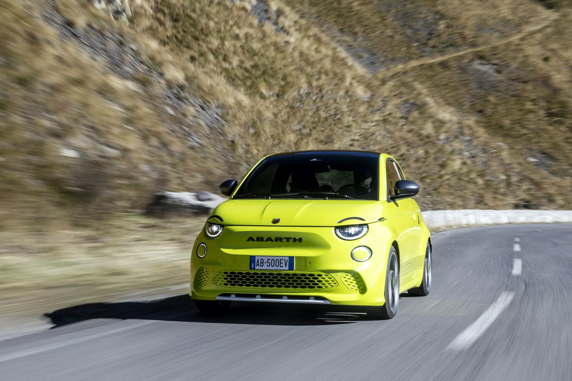 Jasnožlté auto Abarth 500-595-695 2023 druhej generácie zachytené v pohybe na horskej ceste. Obrázok, odfotený z predného trojštvrťového uhla, zobrazuje prednú ľavú stranu vrátane prednej masky, nárazníka, svetlometov, kapoty, čelného skla, dverí vodiča a predného kolesa. Športový hatchback má zväčšené prívody vzduchu, výrazné zliatinové kolesá, nižšiu svetlú výšku, elegantné LED svetlomety a aerodynamicky tvarovanú kapotu. Rozmazanie pohybu naznačuje vysokú rýchlosť s horským pozadím a jasnou modrou oblohou, zdôrazňujúcim dizajn a výkon vozidla.