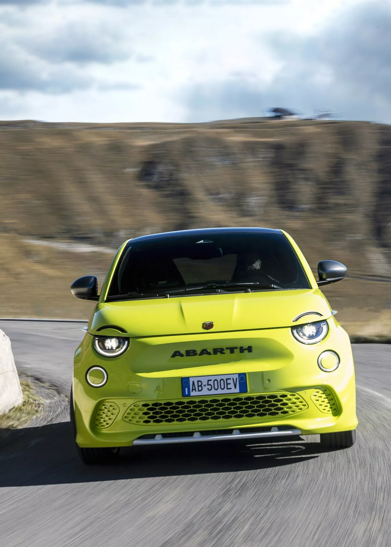 Obrázok Abarth 500-595-695 2023 (2. generácia) kompaktného športového vozidla v pohybe, pohľad zpredu s miernym sklonom doprava, ktorý ukazuje aerodynamický dizajn, pokročilú a LED technológiu osvetlenia, emblém na prednej maske a funkčné prvky nasávania vzduchu, zachytený počas dňa v vidieckom prostredí.