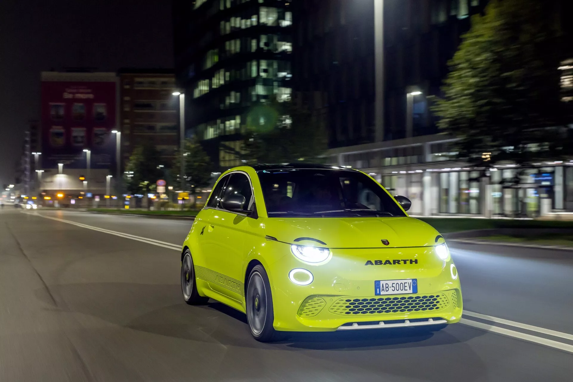 Dynamická nočná snímka živého limetkovozeleného Abarthu 500-595-695 2023 (2. generácia) v pohybe, s dôrazom na prednú a spolujazdcovu stranu z nízkeho čelného uhla so svietiacimi svetlometmi a dennými LED svetlami, športová predná mriežka a zliatinové kolesá, na tmavom mestskom pozadí zdôrazňujúce športový dizajn vozidla a mestské prostredie jazdy.