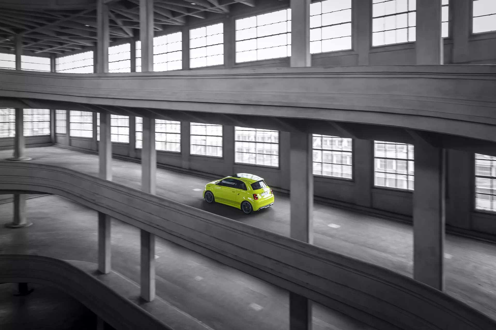 Záber z vysokého uhla zachytávajúci limetkovo zelený hatchback Abarth 500-595-695 2023, ktorý jazdí po zakrivenej betónovej rampe v priemyselnom prostredí, pričom monochromatická sivá architektúra zvýrazňuje dynamický pohyb a moderný aerodynamický dizajn auta.