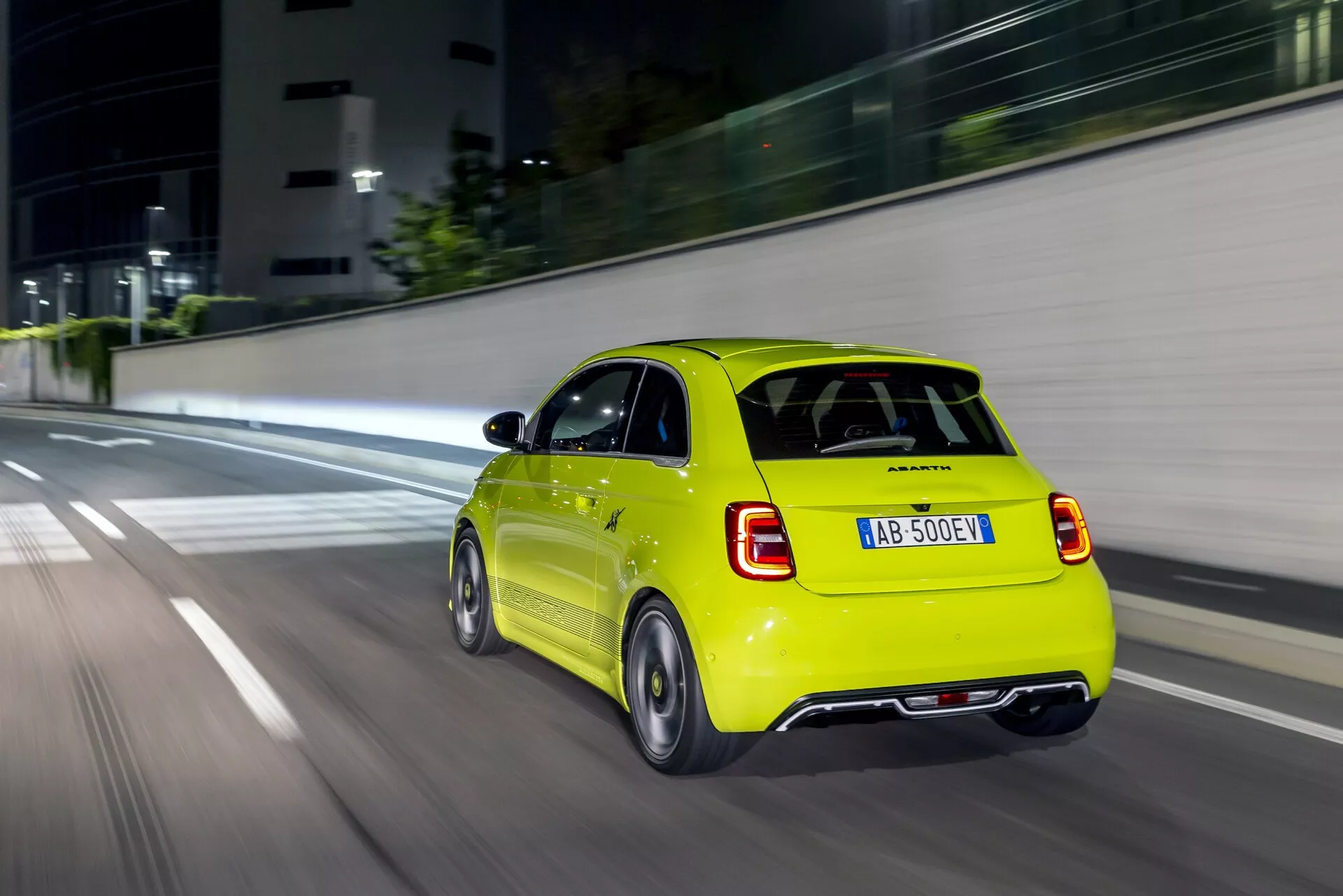 Zadný pohľad na jasne žltý Abarth 500-595-695 2023 (2. generácie) hatchback v pohybe, zachytený z prenasledovacieho uhla mierne vľavo a za vozidlom. Scéna s ľahkými trakmi a rozmazaním pohybu na kolesách a prostredí dáva dojem pohybu v miernej až vyššej rýchlosti. Viditeľné sú zadné svetlá, zadný nárazník, veko batožinového priestoru s centrálnou ešpézetkou a označenie modelu/výrobcu na spodnej pravej strane plus ďalšie označenie vľavo. Osvetlené zadné svetlá naznačujú nízke svetelné podmienky alebo zapojené brzdové svetlá. Záber, pravdepodobne vyhotovený v noci alebo večer v mestskom prostredí s umelým osvetlením a modernými štruktúrami jemne viditeľnými v pozadí.