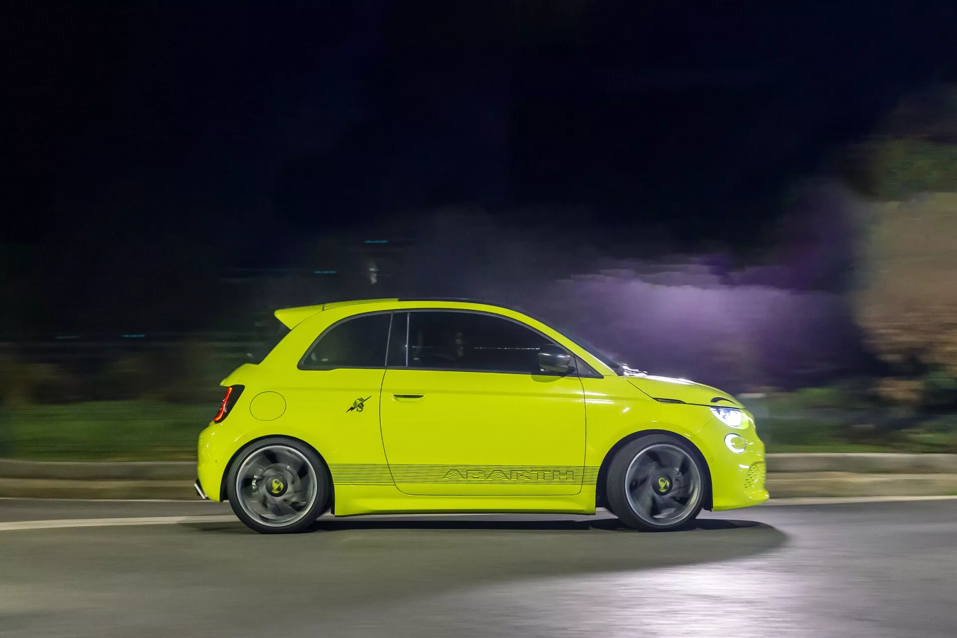 Dynamický bočný pohľad na jasne žlté auto Abarth 500-595-695 2023 (2. generácie) v pohybe v noci, zachytený technikou panorámovania. Auto je ostro zaostrené, zatiaľ čo pozadie je rozmazané, čo vyjadruje rýchlosť. Má dvojdverový dizajn, bočné zrkadlá, kľučky dverí, kolesá, bočné panely a charakteristický zadný spojler. Uličné osvetlenie osvetľuje auto, zvýrazňujúc jeho dizajnové línie a kvalitu laku.