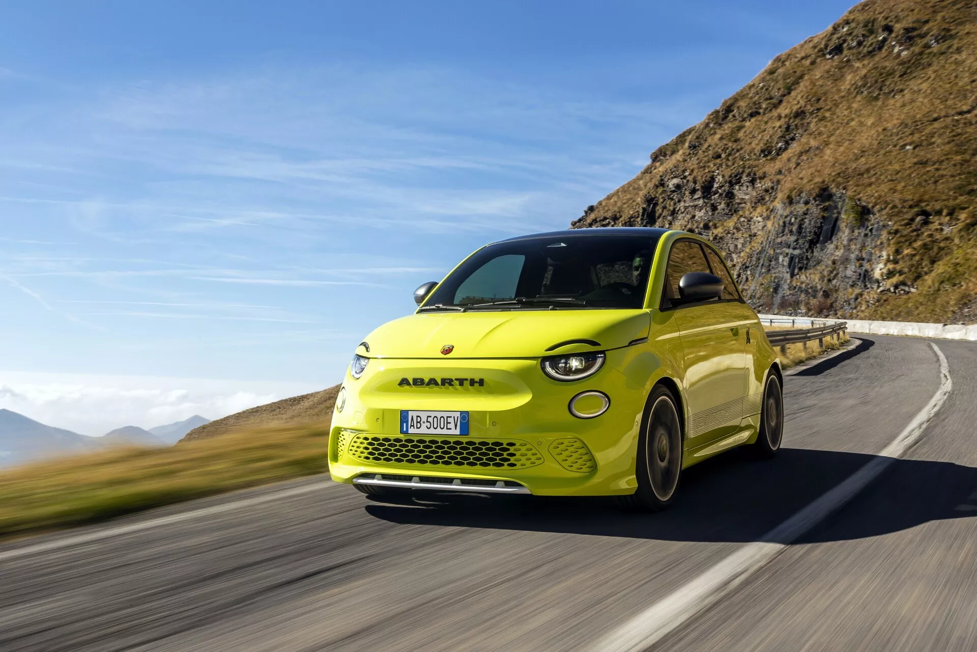 Obrázok jasne žltého auta Abarth 500-595-695 2023 (2. generácia) v pohybe na horskej ceste pod jasnou modrou oblohou. Pohľad z troch štvrtín spredu zdôrazňuje prednú mriežku, svetlomety, bočný profil a športový dizajn. Rozmazanie kolies naznačuje vysokú rýchlosť. 'e' odznak na zadnej strane naznačuje, že ide o elektrický model s viditeľnými LED dennými svetlami a moderným svetelným klastrom.