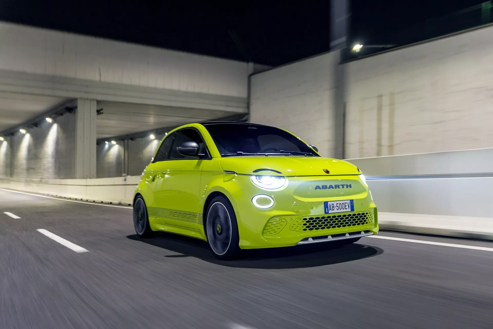 Žiarivo žltý Abarth 500-595-695 2023 (2. generácia) v pohybe, zachytený v nočnom alebo slabo osvetlenom tuneli. Zobrazený z nízkeho trojštvrťového predného pohľadu, zvýrazňujúci prednú pravú stranu vrátane kapoty, blatníka, svetlometu a mriežky. Rozmazanie pohybu naznačuje strednú až vysokú rýchlosť s točiacimi sa kolesami. Dominantné LED svetlomety s výrazným denným osvetlením. Športové hliníkové kolesá a výkonné pneumatiky odrážajú zameranie spoločnosti Abarth na športovosť a výkon.