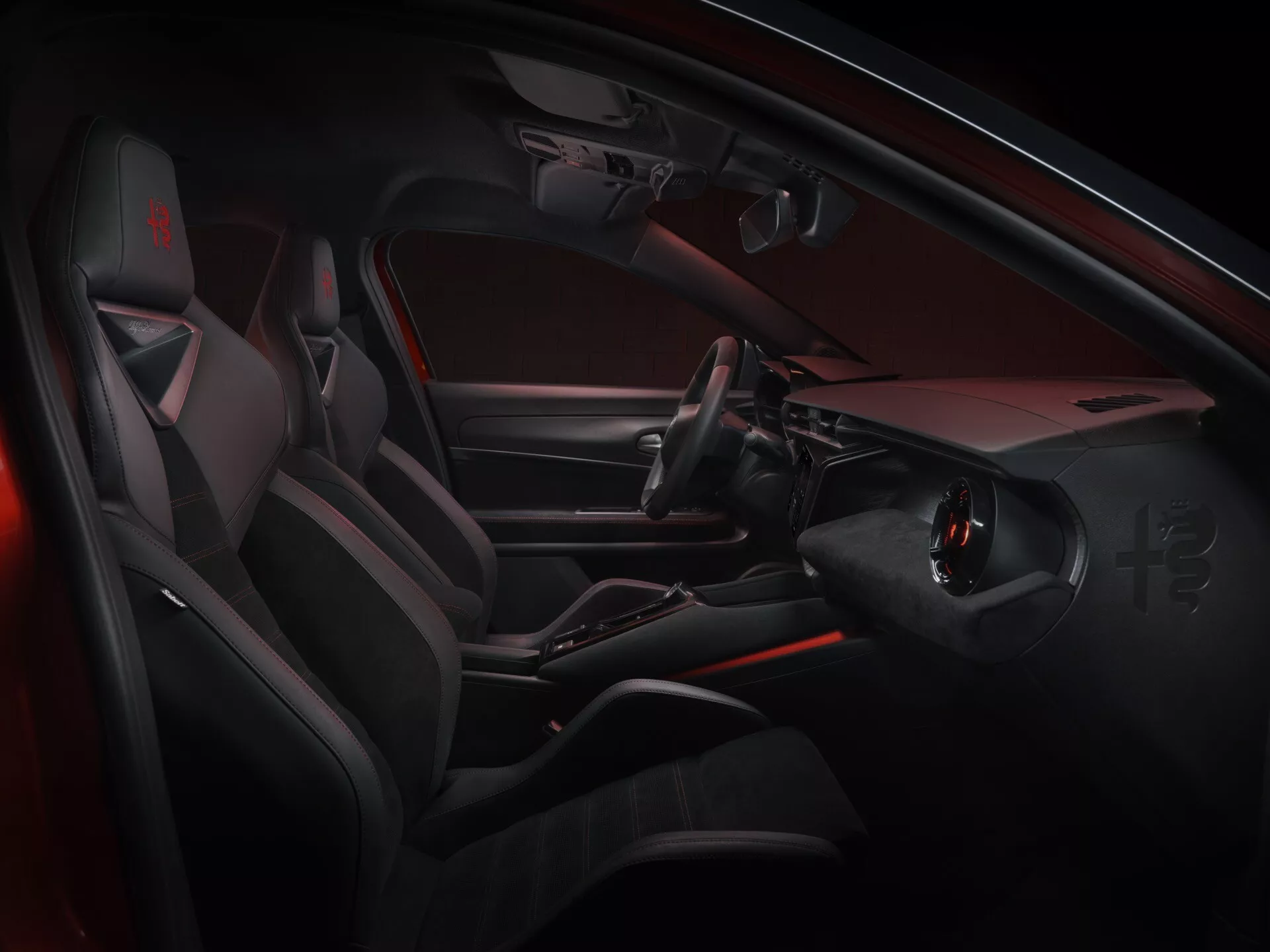 Pohľad do interiéru Alfa Romeo Junior 2024 ukazujúci stranu vodiča s modernými, športovými prvkami dizajnu vrátane volantu s plochým dnom a integrovanými ovládačmi, elegantnou stredovou konzolou bez tradičnej radiacej páky, športovými sedadlami s výraznými bočnými výstuhami a luxusnými materiálmi s akcentmi červeného ambientného osvetlenia