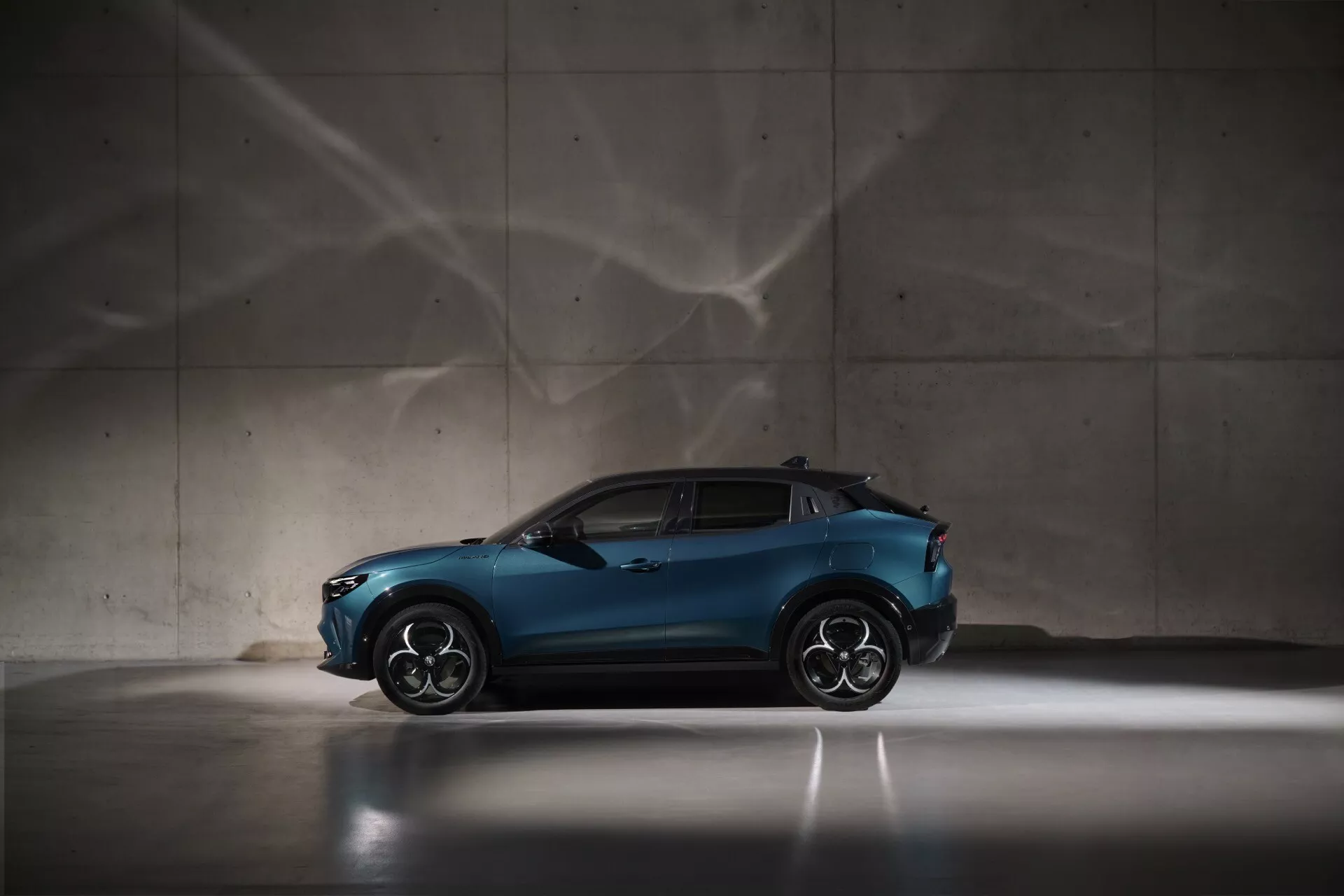 Alfa Romeo Junior 2024 v metalickej modrej, bočný pohľad predstavujúci športový a kompaktný dizajn vozidla crossover, s veľkými zliatinovými kolesami a sklonenou strechou, s dramatickým osvetlením a umeleckými tieňovými vzormi na pozadí