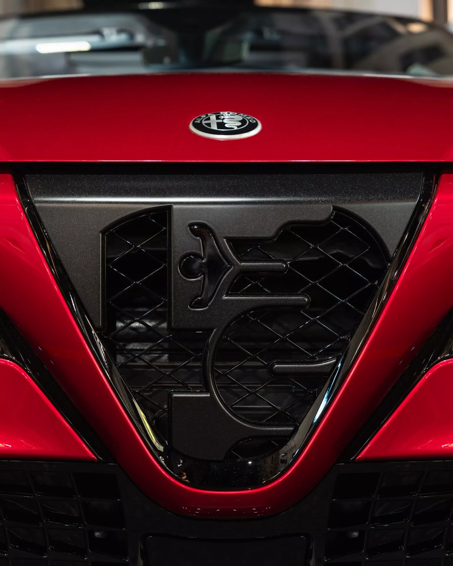 Detailný pohľad na mriežku a odznak výrobcu Alfa-Romeo Junior (966) pre rok 2024, zdôrazňujúci zložitý hexagonálny sieťový dizajn, aerodynamické prvky a lesklý červený povrch