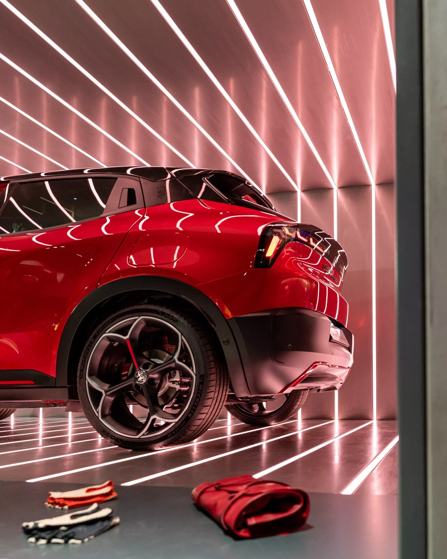 Detailný pohľad na červené Alfa Romeo Junior 2024 s dôrazom na predný štvrťový panel, zdôrazňujúci pokročilý systém predných svetiel, športový dizajn kolesa a odrazivú povrchovú farbu v interiéri výstavnej haly