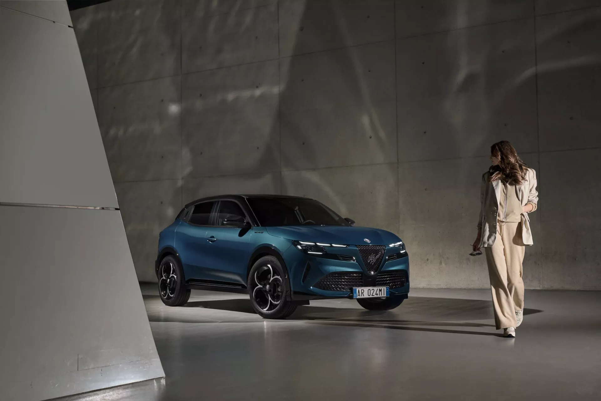 Štýlové SUV Alfa Romeo Junior 2024 predstavené v modernom minimalistickom showroome s dramatickým horným osvetlením zdôrazňujúcim jeho živú modrú farbu, pokročilé LED svetlomety a ikonický dizajn prednej masky, zachytené z trojštvrtovej prednej perspektívy