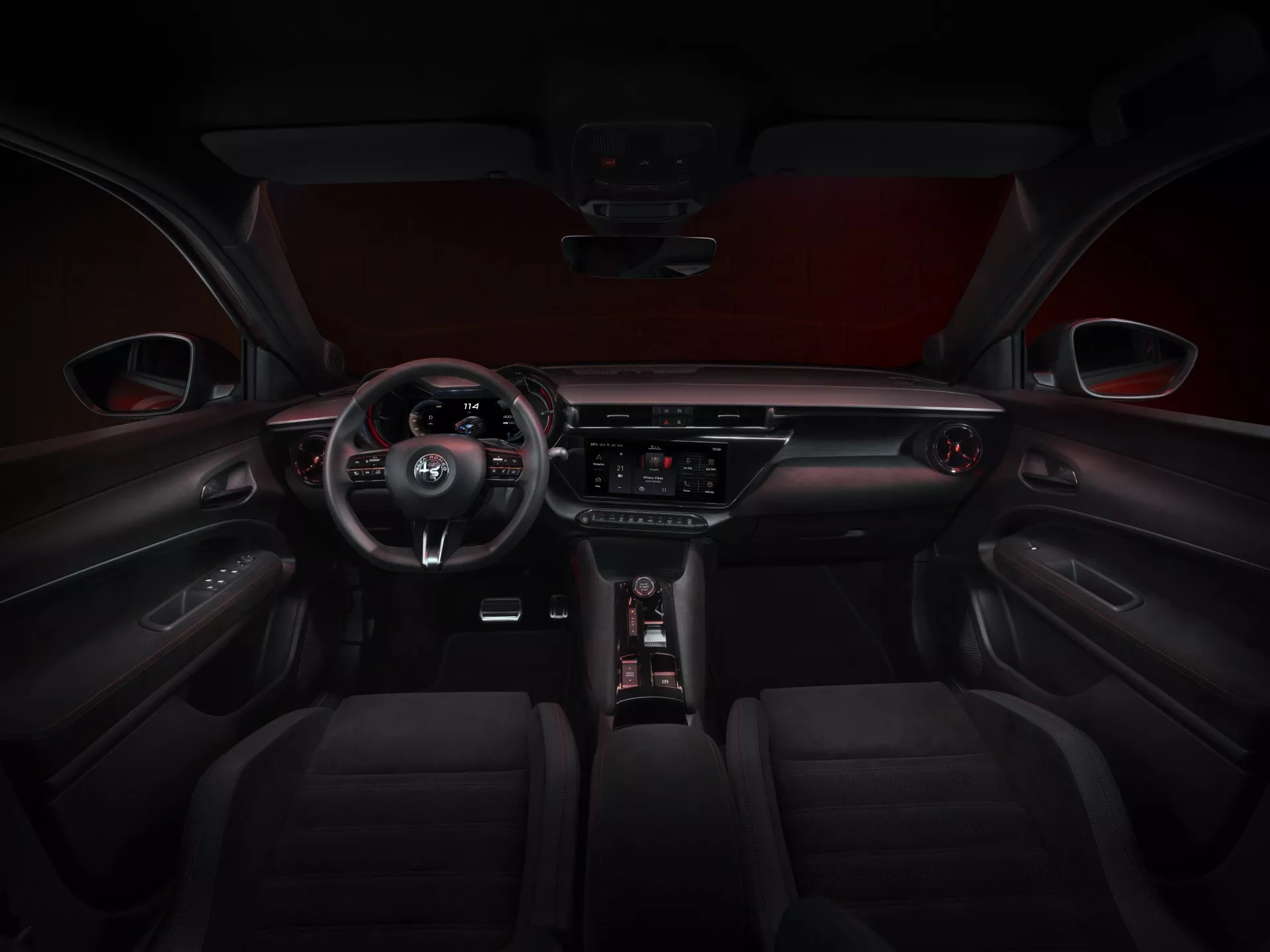 Interiérový pohľad na Alfa-Romeo Junior 2024 (966) ukazujúci tmavú koženú alebo koženkovú čalúnenie s viditeľným šitím na predných sedadlách, trojlúčový volant s integrovanými ovládacími prvkami s emblémom výrobcu, čiastočne viditeľný digitálny prístrojový panel, centrálny dotykový informačno-zábavný systém s tlačidlami pre riadenie klimatizácie a ďalšie ovládacie prvky vozidla pod ním, elegantnú radiacu páku po boku intuitívneho ovládacieho gombíka, doplnený kontrastným červeným čalúnením strechy a dverovými panelmi s integrovanými ovládacími prvkami a šitím.