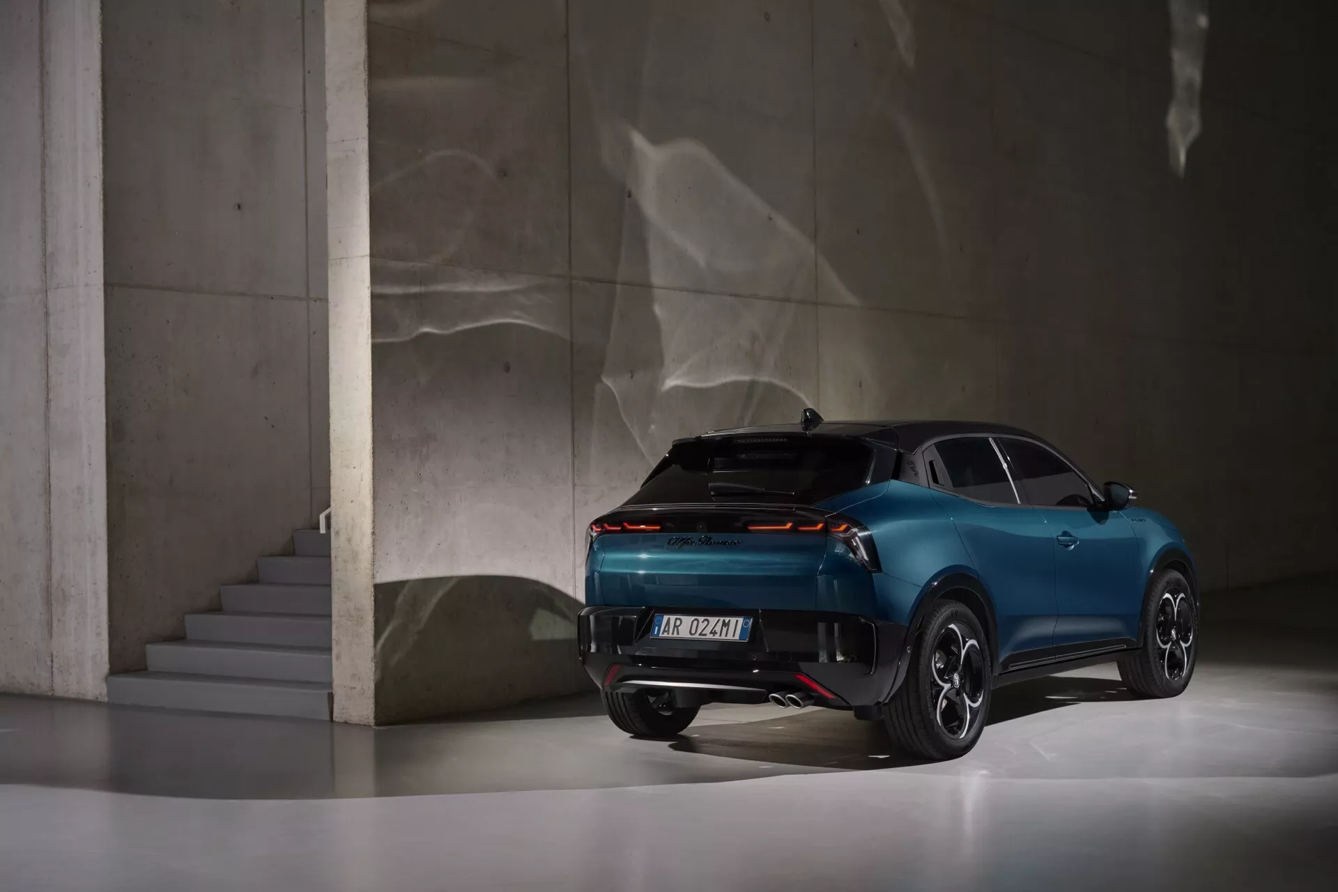 Alfa Romeo Junior 2024 zobrazené z zadného trojštvrťového pohľadu ukazuje jeho moderný dizajn, zadné svetlá pokrývajúce takmer celú šírku vozidla, športovo-kupéovú siluetu so štyrmi dverami a luxusné prvky, umiestnené v elegantnom interiéri s odrazivou podlahou a betónovými konštrukciami.