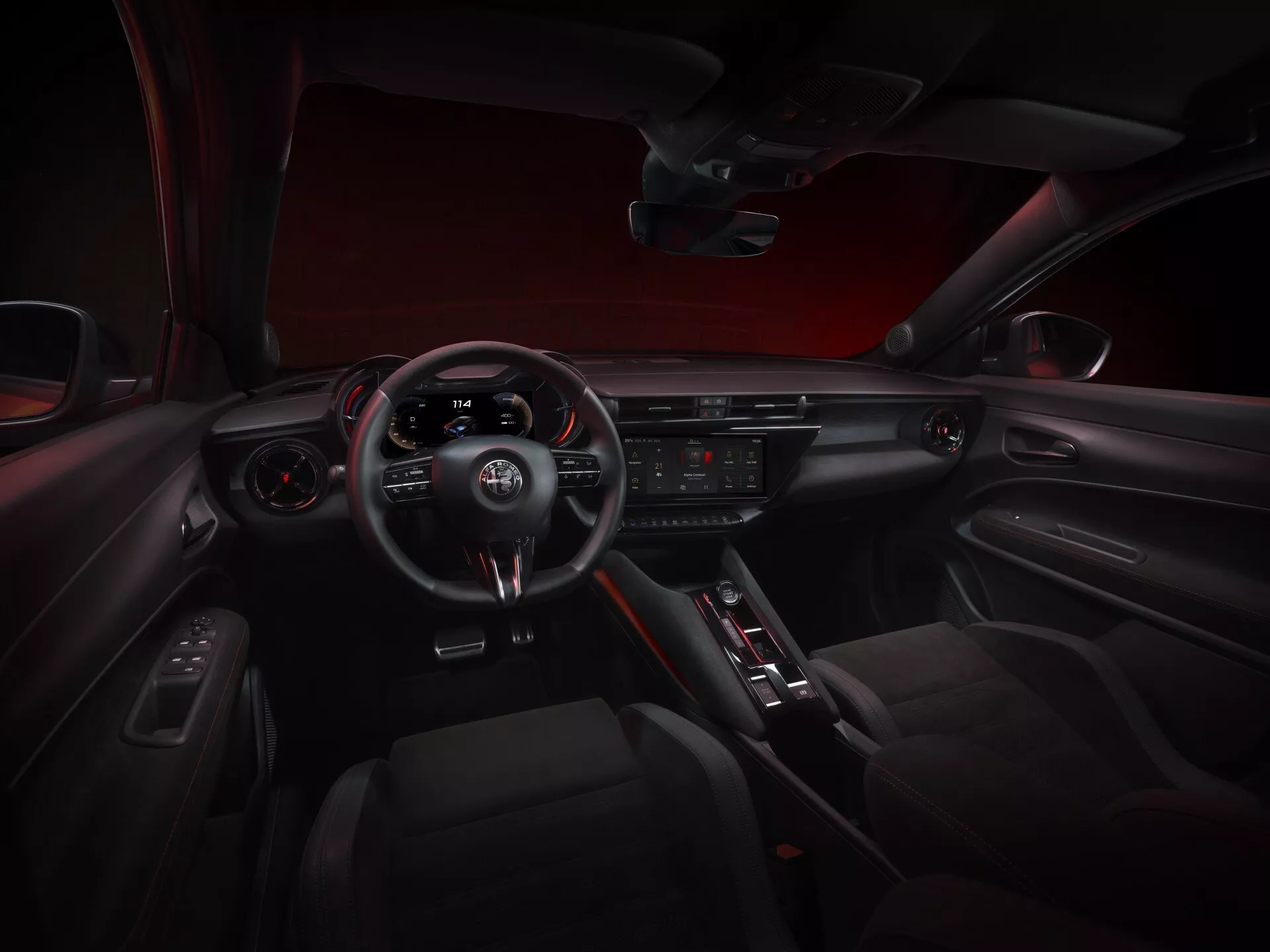 Interiérový pohľad na kokpit Alfa-Romeo Junior 2024 z centrálneho zadného sedadla, zobrazujúci prístrojovú dosku, športový volant so troma ramenami s integrovanými ovládačmi a radením na volante, prístrojový panel, centrálny displej infotainment systému, tlačidlá klimatizácie, volič automatickej prevodovky, panel dverí s elektrickými ovládaniami a športovo elegantnú atmosféru s červeným akcentujúcim osvetlením