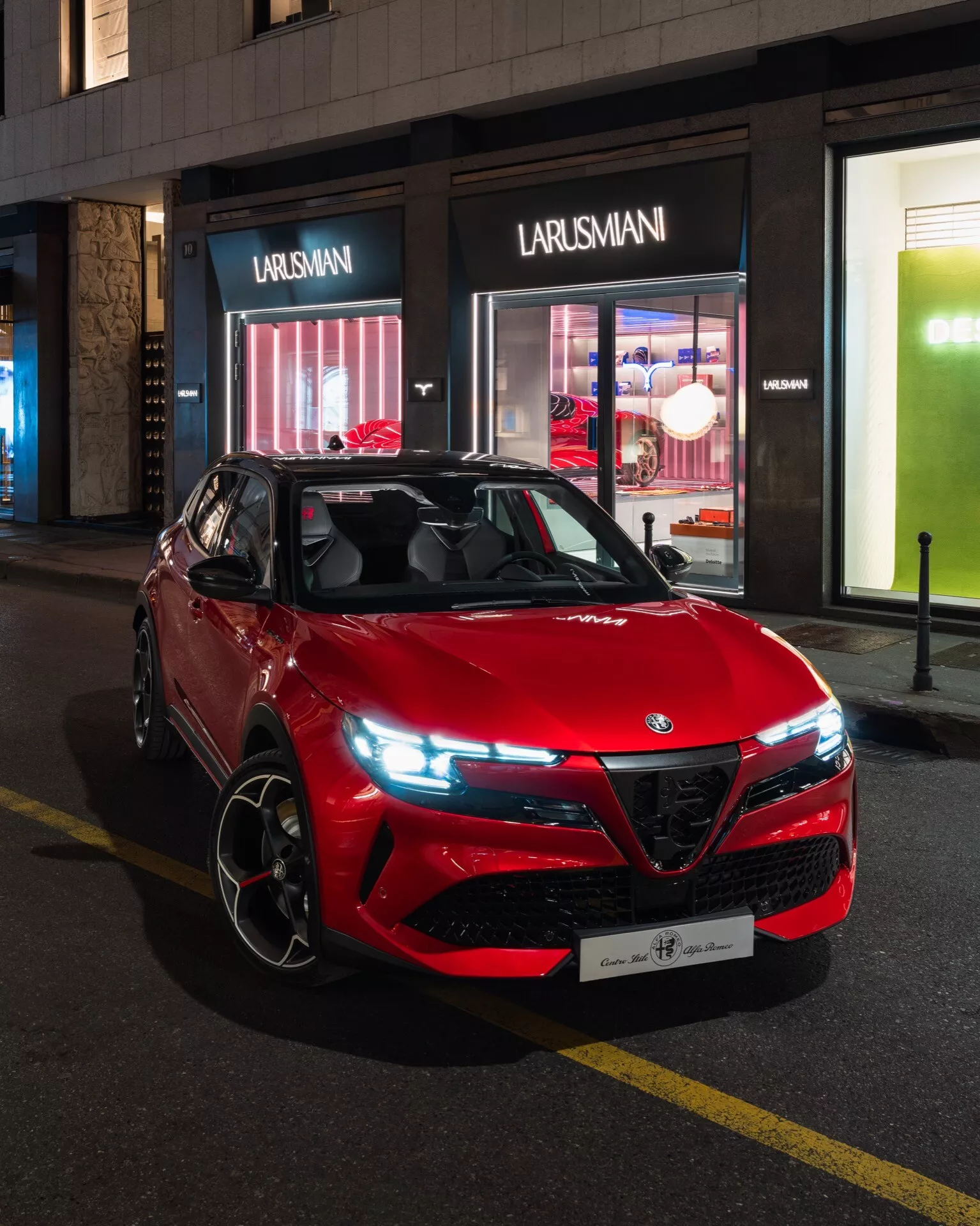 Červené športové utility vozidlo Alfa-Romeo Junior 2024 zaparkované na mestskom ulici v noci, predvádzajúce svoj športový a agresívny dizajn s osobitou mriežkou, modernými LED svetlometmi a výkonnostne orientovanými zliatinovými kolesami