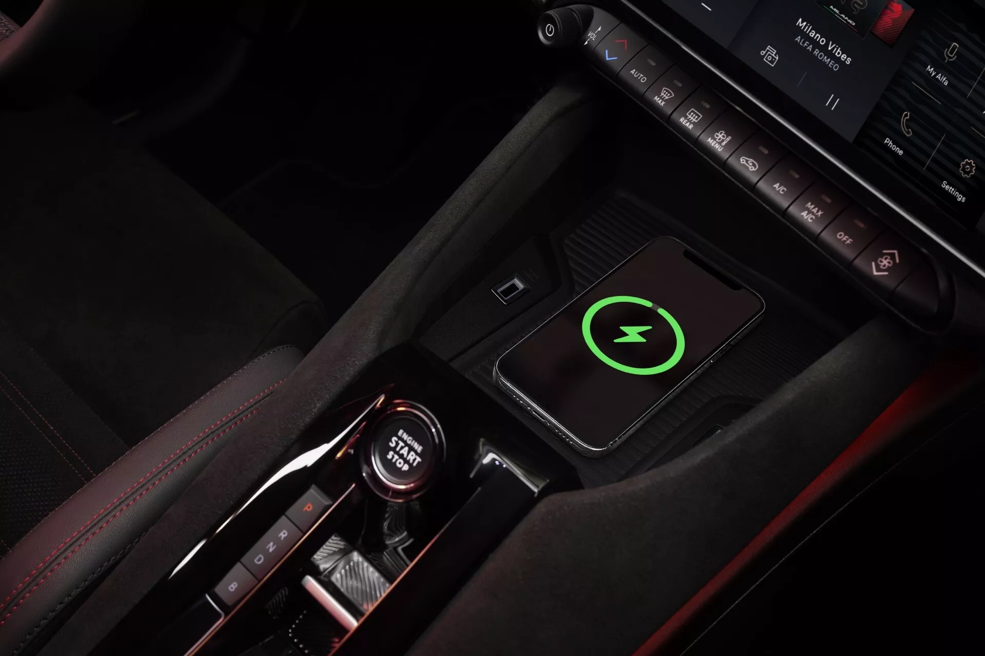 Zhora pohľad na stredovú konzolu Alfa-Romeo Junior 2024 zdôrazňujúci červené švy, štýlovú páku prevodovky s indikátormi automatickej prevodovky, bezdrôtovú nabíjaciu podložku so zeleným symbolom blesku, ovládacie tlačidlá, ambientné osvetlenie, držiak na poháre s červenými akcentmi, zvýrazňujúci interiér zameraný na luxus a technológiu