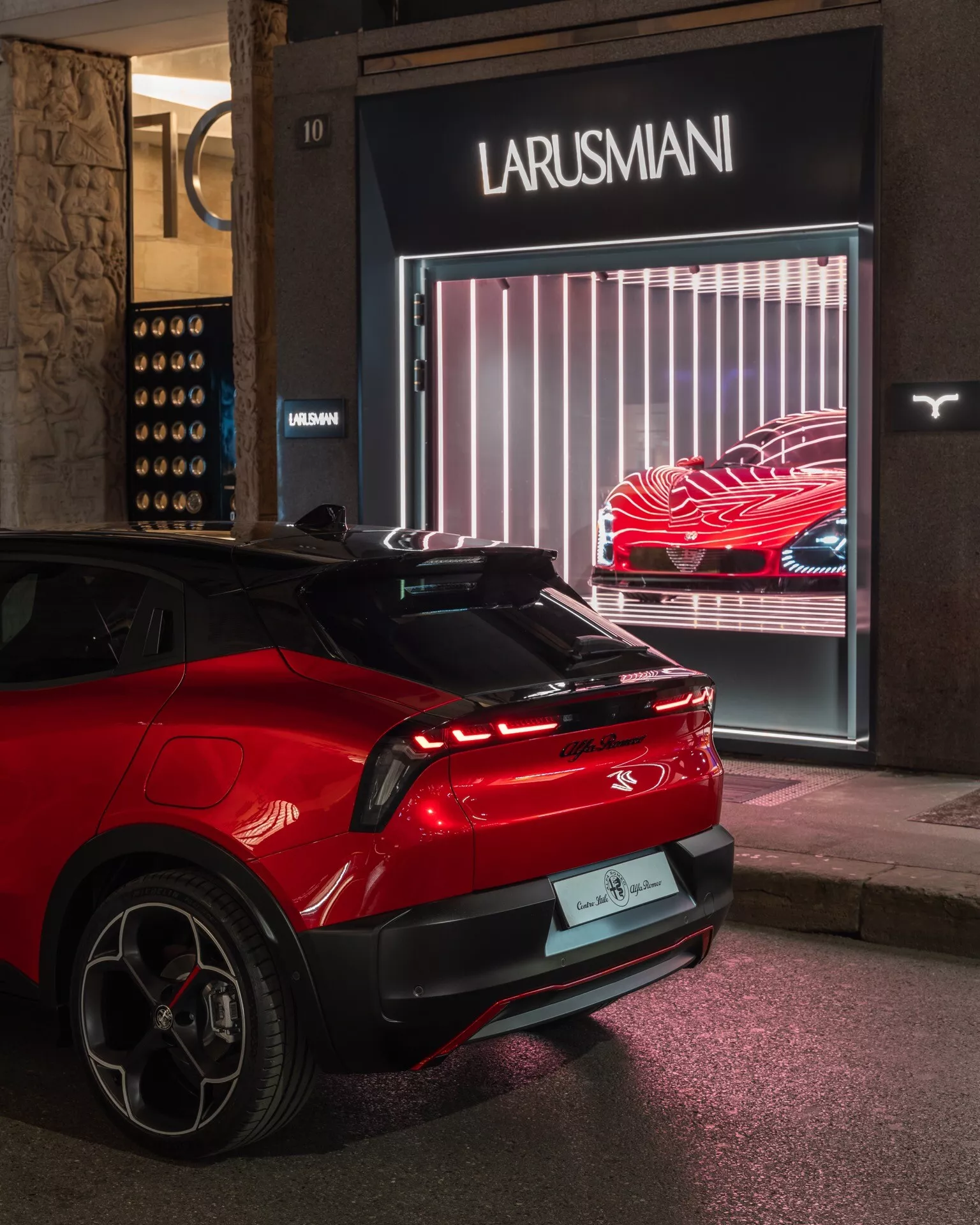 Zadný trojštvrťový pohľad na moderné červené Alfa Romeo Junior 2024 zaparkované v noci pred obchodom Larusmani, ukazujúce LED zadné svetlá, zadné čelné sklo so stieračom, strešný spojler, zliatinové kolesá s nízkoprofilovými pneumatikami a naznačujúce elektrickú alebo hybridnú technológiu, v mestskom osvetlení.