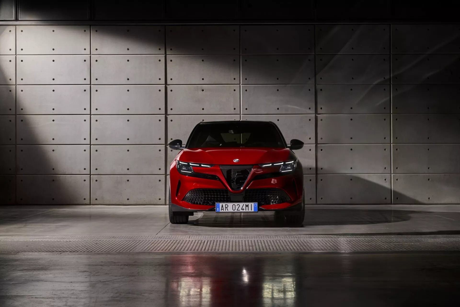 Pohľad spredu na Alfa Romeo Junior 2024, zachytený z nízkeho uhla na kovovej mriežkovej podlahe, s LED svetlometmi, výraznou mriežkou s odznakom výrobcu a štýlovým dizajnom nárazníka, na pozadí betónovej steny s kruhovými vydutinami pod dramatickým osvetlením.