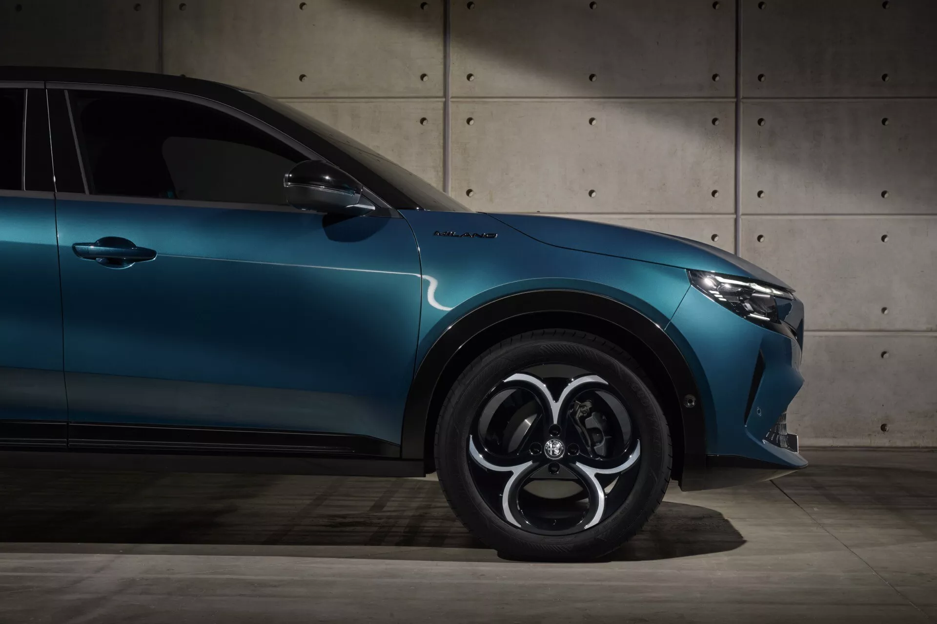 Záber z nízkeho uhla na Alfa-Romeo Junior (966) 2024 zdôrazňujúci jeho bočný profil, s odznakom TurboS, vysokoúčinnými brzdami, komplikovaným dizajnom disku a elegantnou kovovou modrou farbou