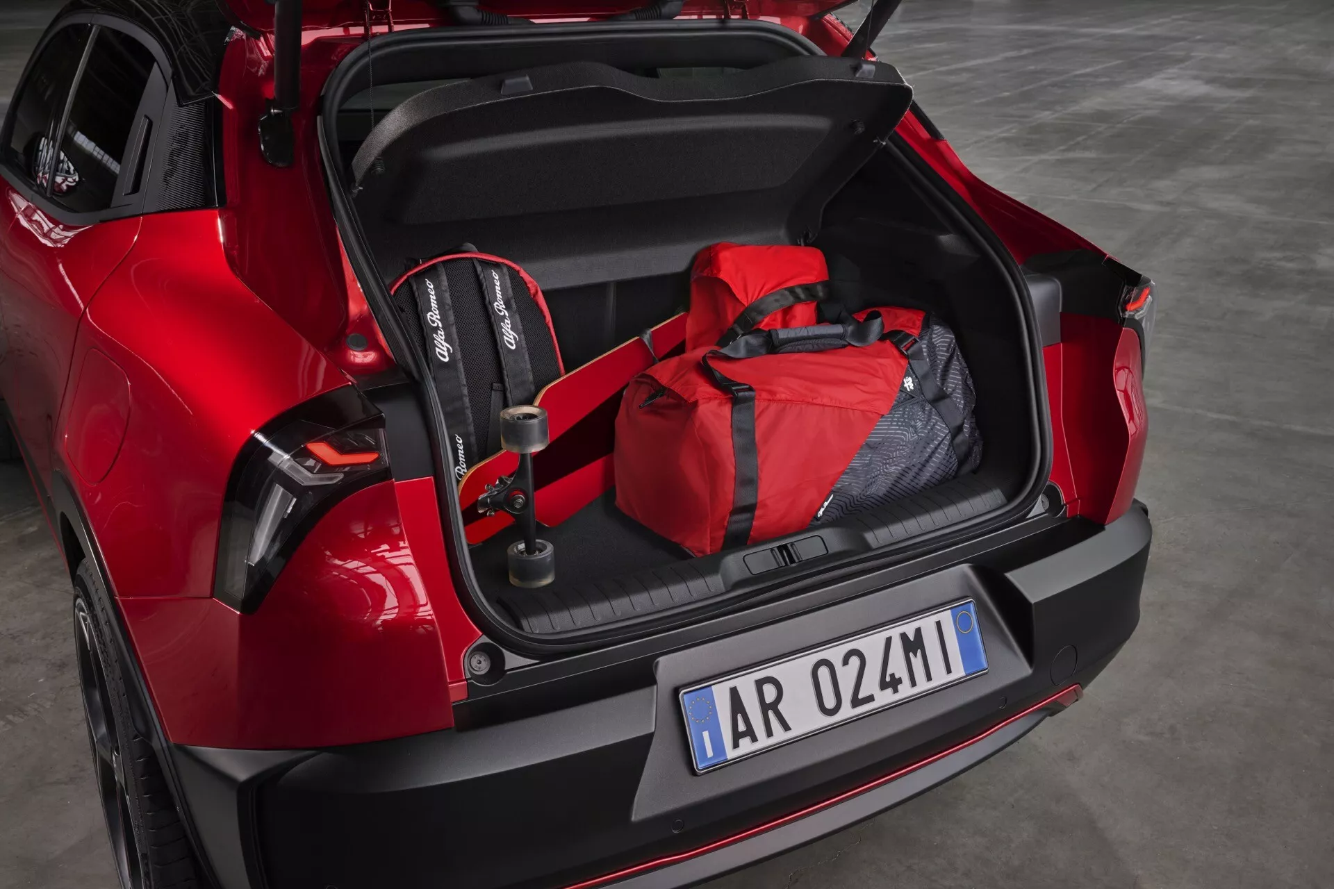 Červený model Alfa-Romeo Junior 2024 s otvoreným kufrom odhaľujúcim priestranný interiér, detskú autosedačku a červenú športovú tašku, s športovým zadným dizajnom a modernými LED zadnými svetlami, čo demonštruje potenciál pre rodinnú priateľskosť a technológiu asistencie pri parkovaní