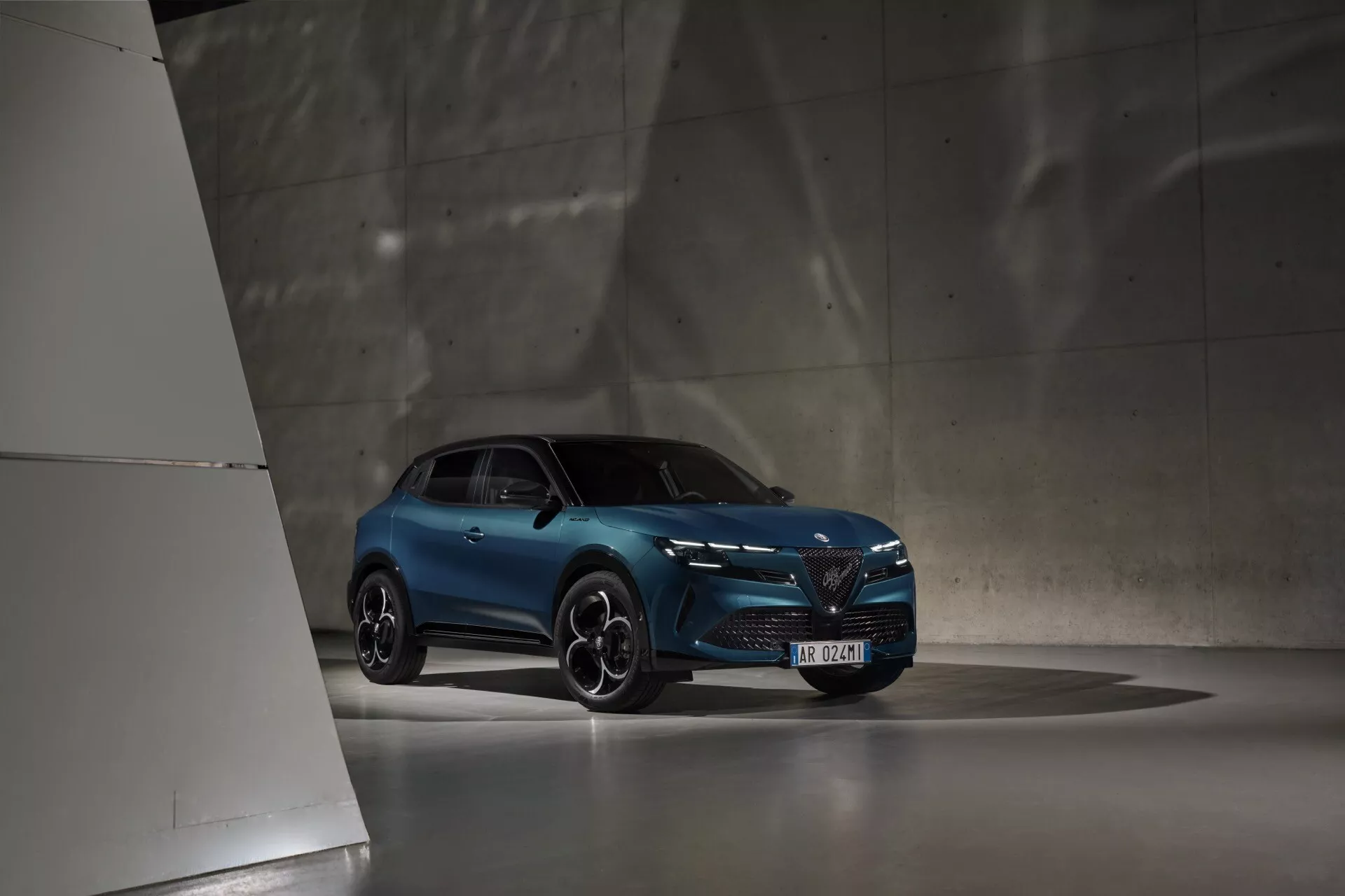 Modrý Alfa Romeo Junior z roku 2024 v slabom osvetlení interiéru, zachytený z nízkeho uhla zvýrazňujúceho jeho moderný dizajn a športový postoj, s LED svetlometmi, výraznou prednou mriežkou a športovými zliatinovými kolesami.