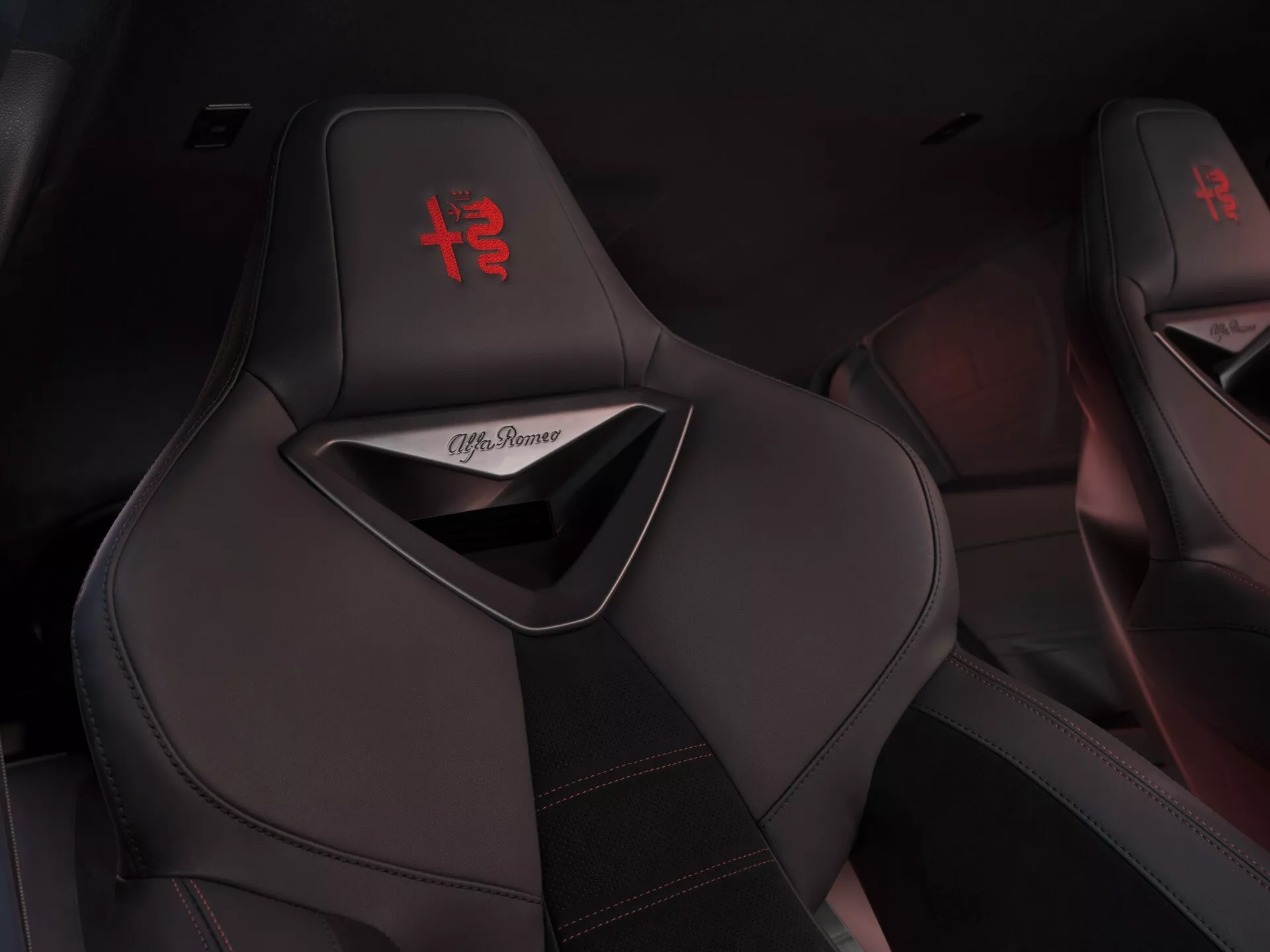 Detailný pohľad na čierne športové sedadlá Alfa Romeo Junior 2024 s vyšitým červeným logom '+' na opierke hlavy a červeným kontrastným prešívaním, ktoré ukazuje luxusný a športový dizajn interiéru