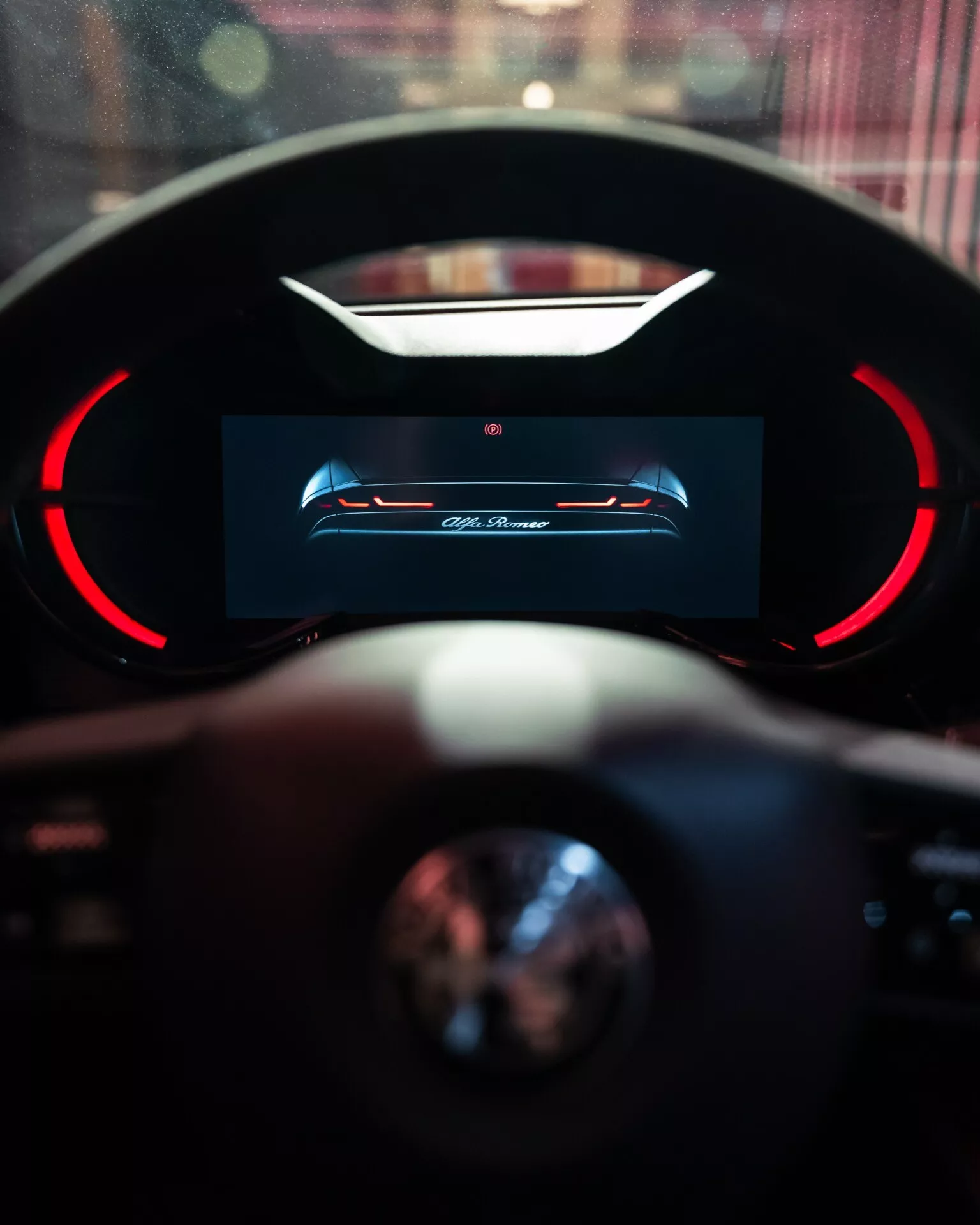 Interiérový pohľad na Alfa-Romeo Junior 2024 s dôrazom na digitálny prístrojový panel vysokého rozlíšenia vodiča s červenými akcentmi a čiastočným pohľadom na volant s rozostreným pozadím naznačujúcim luxusné prostredie