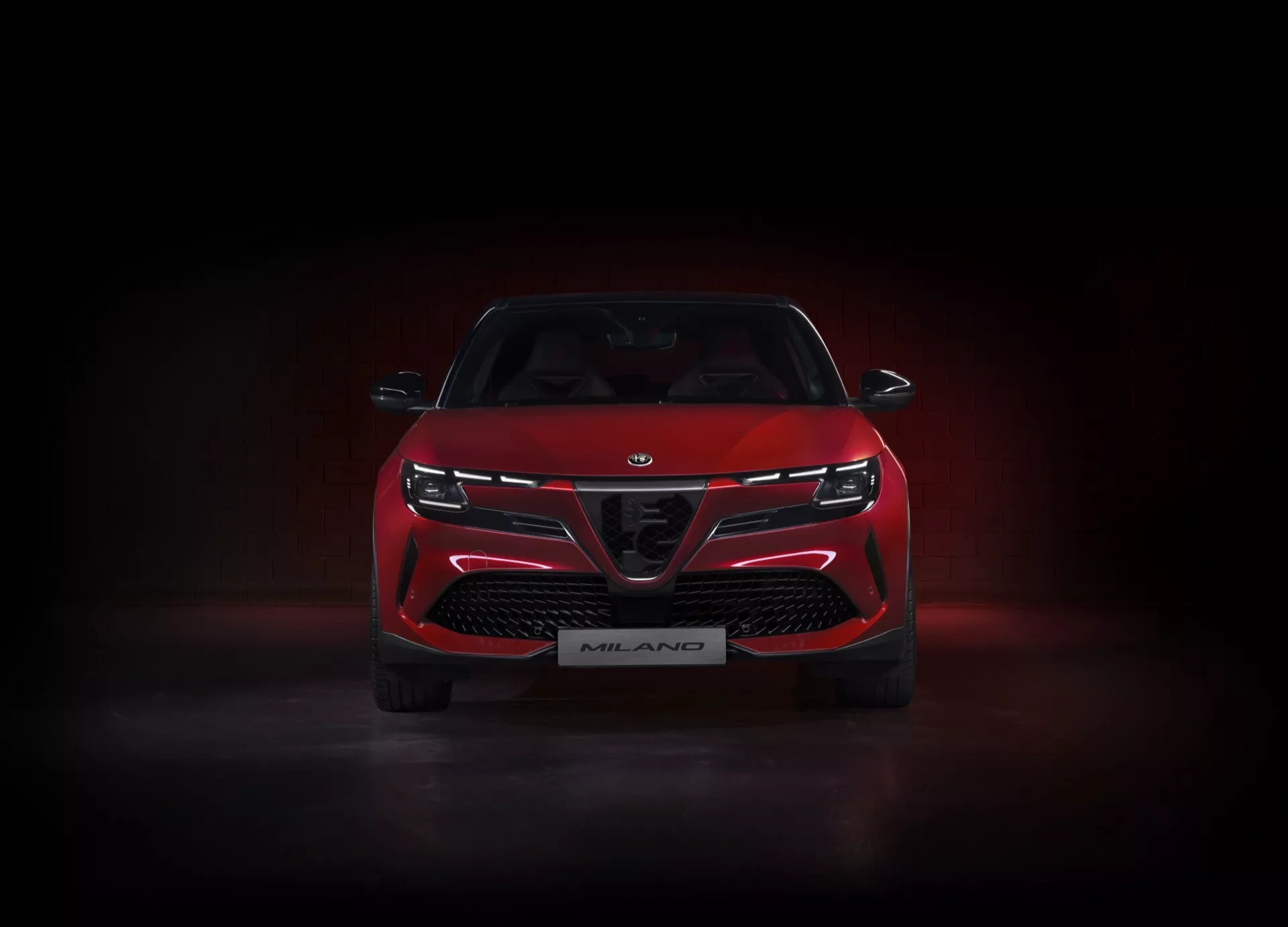 Predný pohľad na červený Alfa-Romeo Junior 2024 (966), osvetlený v štúdiu na tmavom pozadí s jemným spodným svietením, ktorý predstavuje jeho charakteristickú mriežku s veľkými vzduchovými otvormi, elegantné úzke LED svetlomety spojené svetelným pásom, a odznak edície Milano, zdôrazňujúci jeho športové a agresívne dizajnové funkcie.