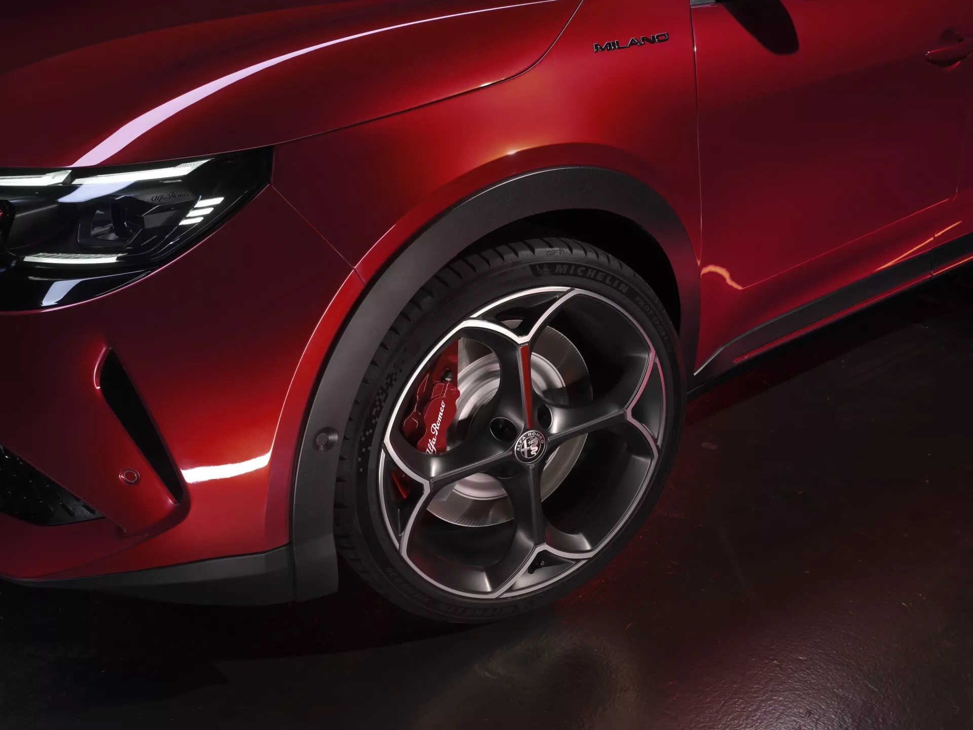Detailný záber na predné ľavé koleso a blatník Alfa Romeo Junior 2024, ukazujúci viacero drátové zliatinové disky, lesklú červenú farbu a červený brzdový strmeň pre prémiový a výkonom orientovaný vzhľad