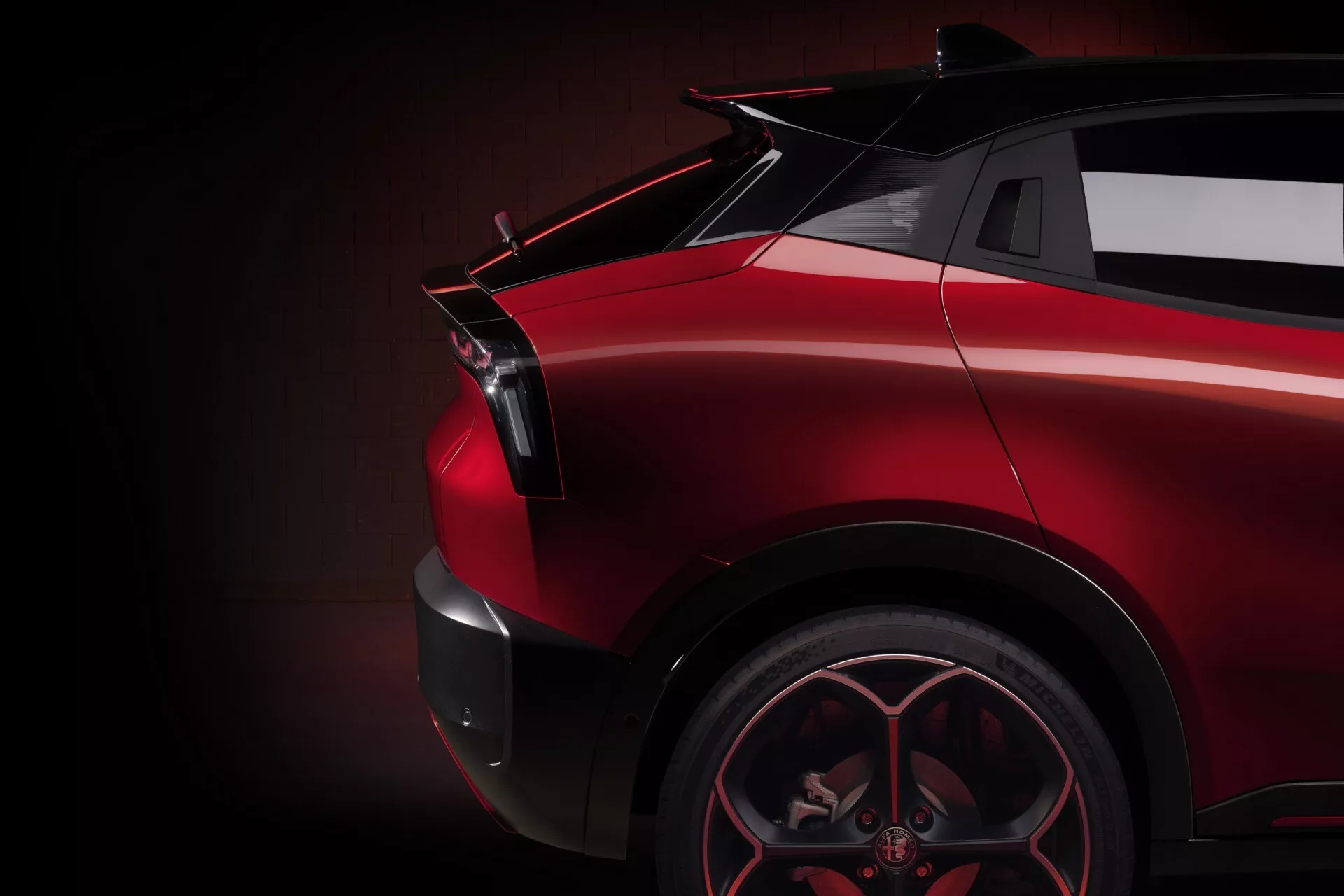 Detailný bočný pohľad na Alfa Romeo Junior 2024 zobrazujúci zadnú štvrtinu na strane spolujazdca so zameraním na športové zliatinové koleso, uhlový zadný blatník, tvarované LED zadné svetlo, lesklý červený lak a možný spojler proti tmavému, jemne osvetlenému pozadiu zdôrazňujúcemu dynamické a luxusné dizajnové prvky vozidla.
