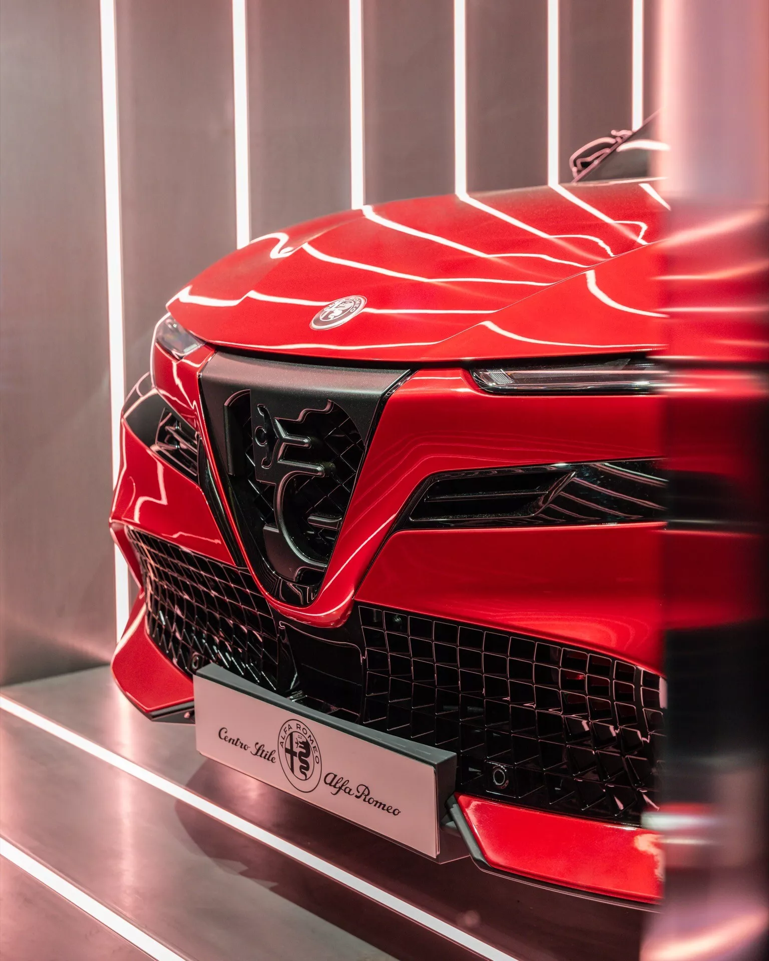 Detailný obrázok prednej ľavej časti Alfa-Romeo Junior 2024 s živou metalickou červenou farbou, ukazujúci jeho agresívny dizajn, ostré uhly, tvarovaný povrch, výrazný emblém, veľkú úhlovú mriežku, elegantné denné svietenie a možné vysokovýkonné vzduchové vstupy, na pozadí jednoduchého interiéru pravdepodobne showroomu.
