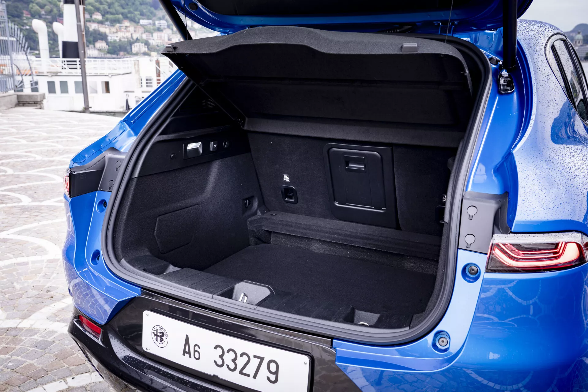 Detailný uhlový pohľad na zadnú časť modrého Alfa Romeo Tonale 2022 s otvoreným kufrom s kobercovým interiérom, integrovanými priehradkami a elektrickou zásuvkou, vedľa viditeľného zadného nárazníka s parkovacími senzormi a modernými LED zadnými svetlami.