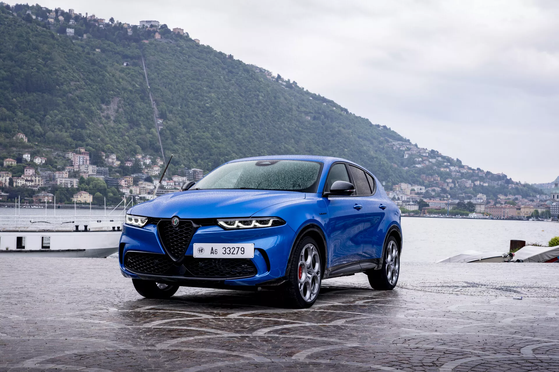 Modré Alfa-Romeo Tonale z roku 2022 zaparkované na mokrom nábreží, predvádzajúce svoju športovú prednú mriežku, LED svetlomety a štíhly profil na pozadí pobrežného mesta