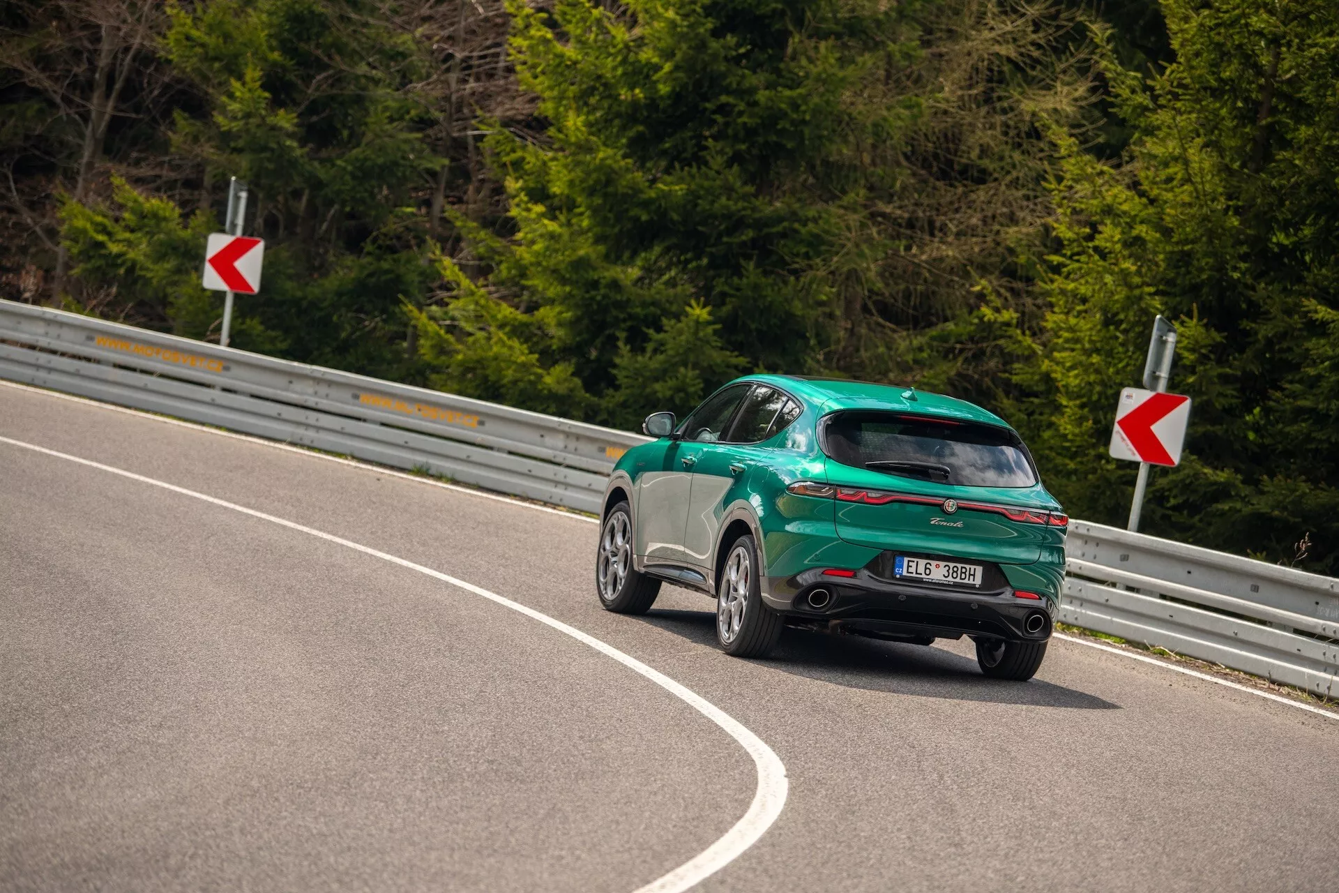 Alfa Romeo Tonale 2022 zachytené z trojštvrťového zadného uhla na zakrivenej ceste, zdôrazňujúce športový dizajn s karosériou hatchback, lesklým povrchom a aerodynamickými funkcie vrátane zadného spojlera, diskových kolies a LED zadných svetiel.
