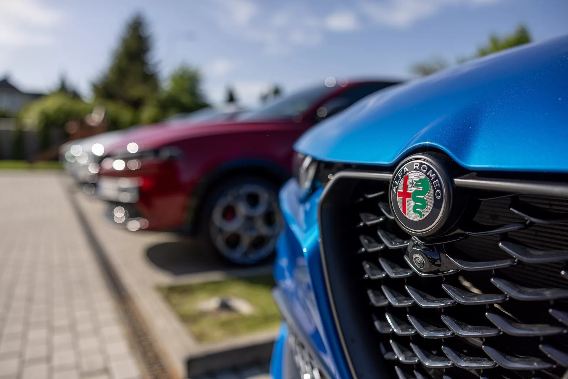 Detailný pohľad na prednú mriežku modrého Alfa Romeo Tonale 2022 so zameraním na emblem, obsahujúci vertikálne lamely a rozmazané pozadie iných vozidiel