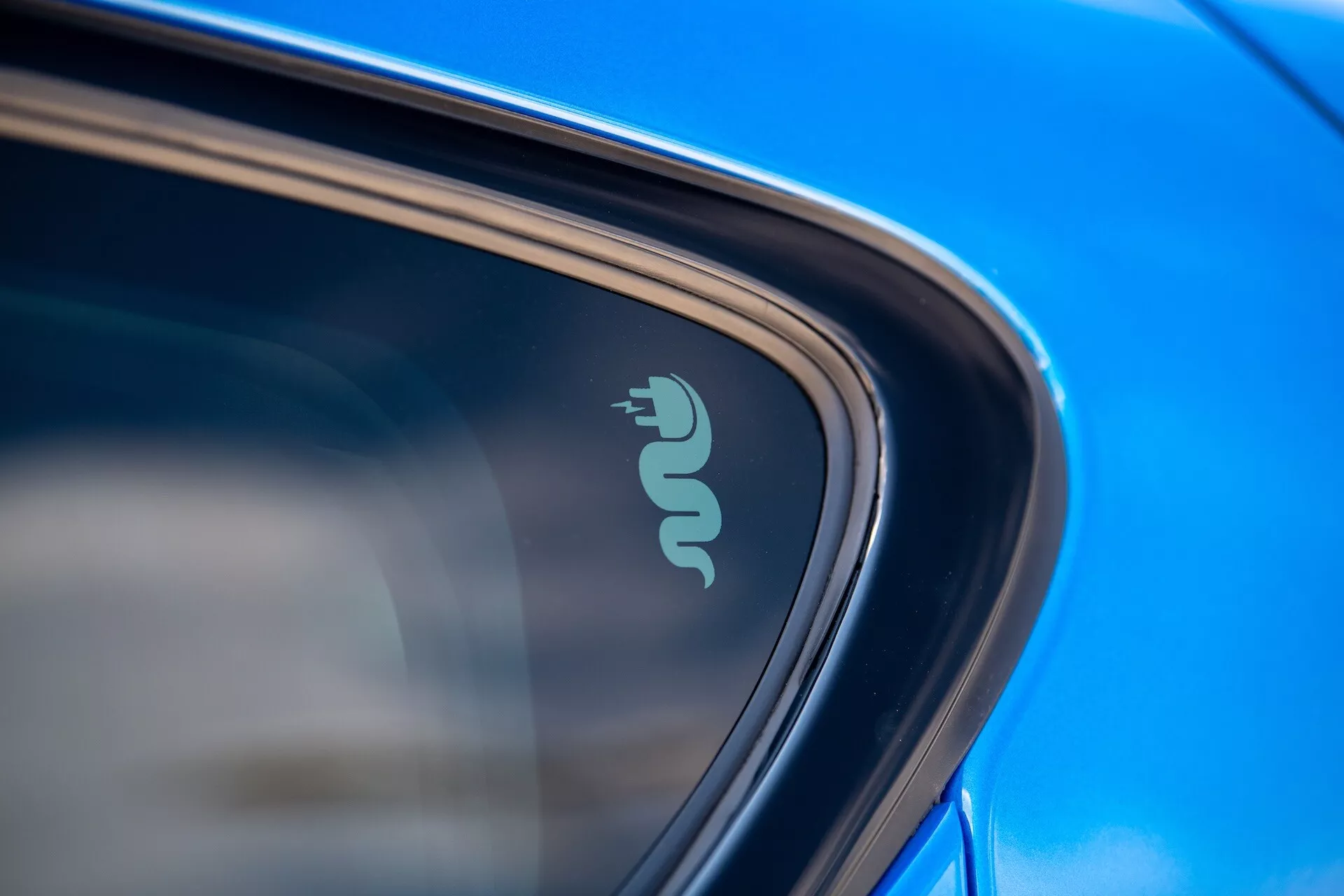 Detailný zbližný záber na technologický znak Alfa Romeo Tonale 2022 naznačujúci elektrickú alebo hybridnú schopnosť blízko okna auta, zvýraznený odrazivou modrou farbou a elegantným dizajnom na prirodzenom dennom svetle