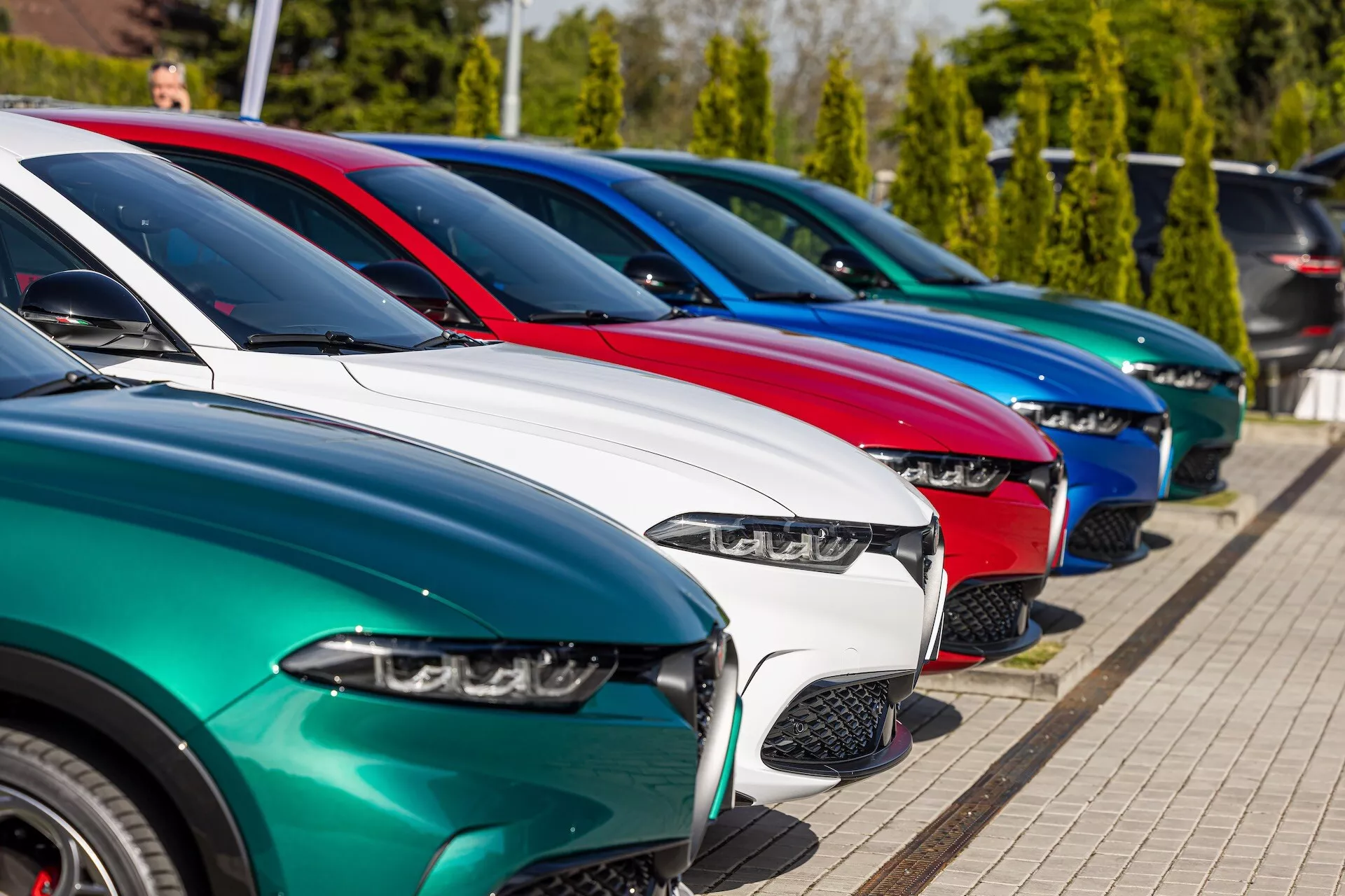 Rad vozidiel Alfa Romeo Tonale 2022 zaparkovaných diagonálne so zobrazením z predného trojštvrtečného uhla zvýrazňujúci LED svetlomety, rôzne dizajny mriežok, lesklé lakové povrchy a zliatinové kolesá na pozadí jasného slnečného svitu a zelene