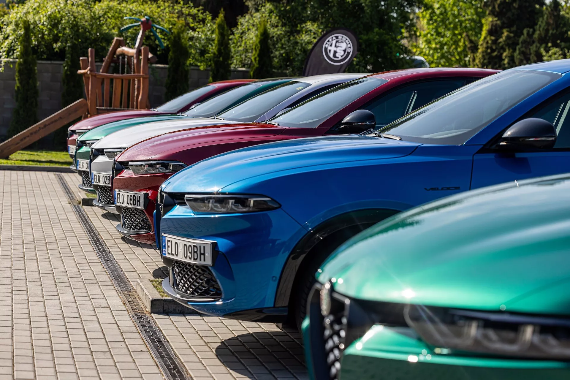 Rad Alfa-Romeo Tonale z roku 2022 v živých farbách ukazuje luxusný dizajn s pokročilými LED svetlami, parkované v diagonálnej formácii so zameraním na prednú ľavú stranu z nízkeho až stredného uhla na spevnenej ploche vedľa trávnatého okraja, možno u dealera alebo na udalosti pre nadšencov automobilov