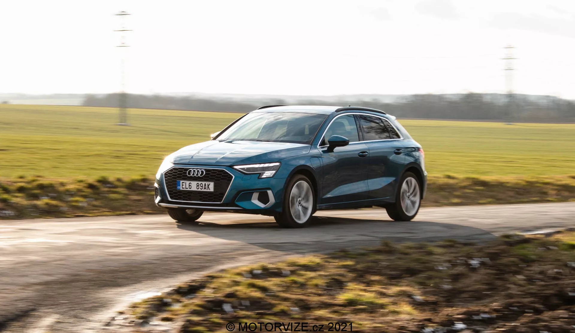 Dynamický záber moderného hatchbacku, pravdepodobne Audi A3 Sportback z roku 2020, zachytený v pohybe. Obrázok je urobený z mierne nízkeho uhla, zdôrazňujúci výšku auta a vytvárajúci dynamický pocit. Ukazuje trojštvrťový predný pohľad na vozidlo pohybujúce sa sprava doľava. Charakteristické dizajnové prvky zahŕňajú elegantné LED svetlá, veľkú prednú mriežku, veľké zliatinové kolesá, aerodynamické bočné zrkadlá s potenciálnymi integrovanými smerovkami a metalický lak s lemovacími líniami. Pozadie je rozmazané, čo zvyšuje pocit rýchlosti, zatiaľ čo samotné auto zostáva v ostrom zaostrení, čím zvýrazňuje svoj moderný dizajn a prémiový vzhľad.