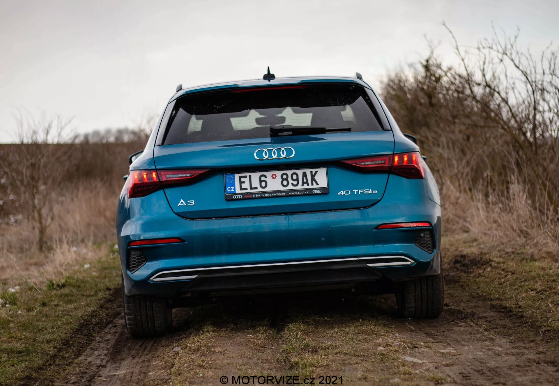 Detailný pohľad na zadnú časť žiarivo modrého Audi A3 Sportback 40 TFSI z roku 2020, zvýrazňujúci jeho pokročilé dizajnové prvky vrátane horizontálnych LED zadných svetiel, dvojitých výfukových koncoviek, znaku TFSI s turbodúchadlom, integrovaného spojlera a žraločej antény, fotografovaný z nízkeho uhla na poľnej ceste s otvoreným poľom a zamračenou oblohou v pozadí.