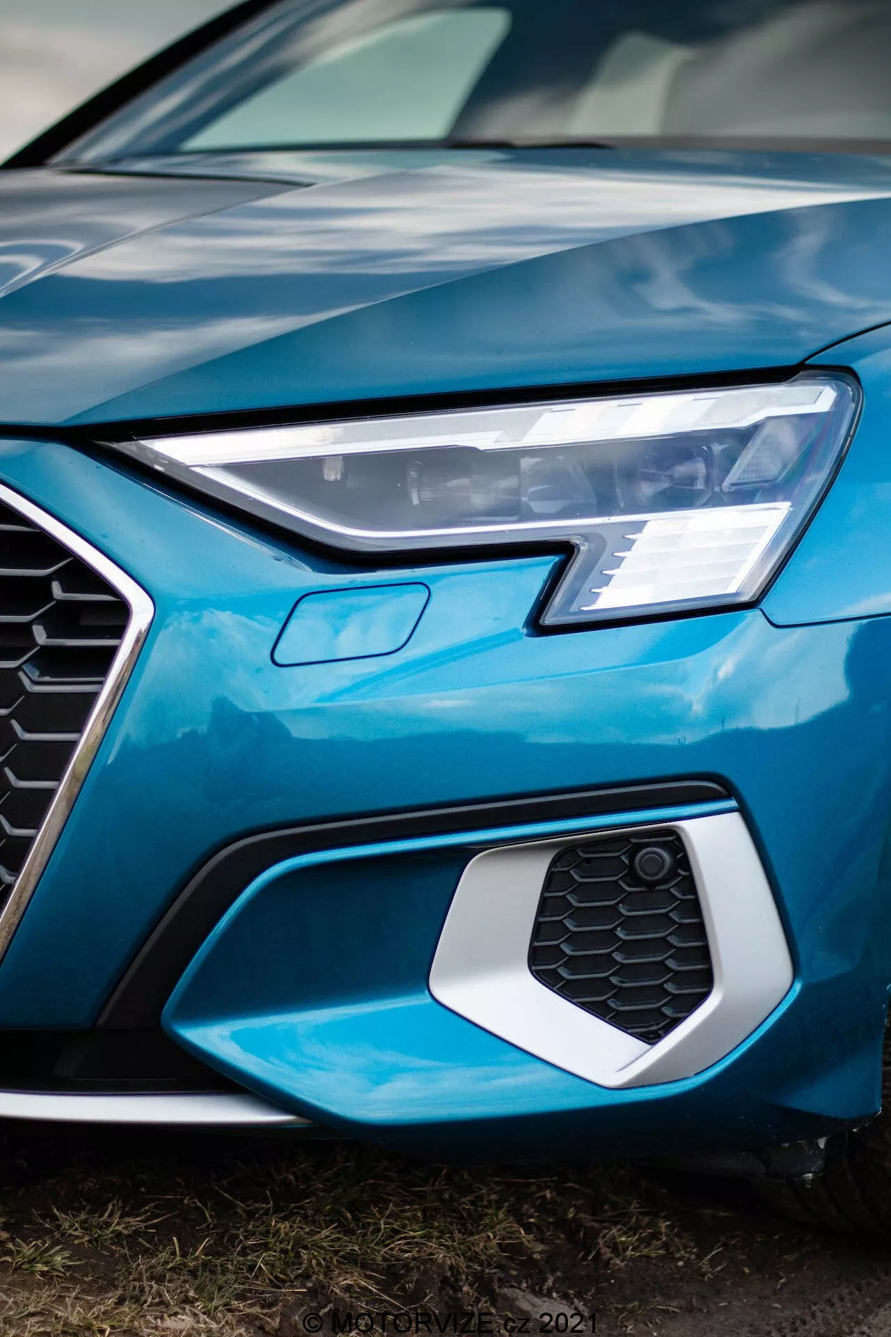 Detailný záber z nízkeho uhla na prednú ľavú časť 2020 Audi A3 Sportback, zachytávajúci ostrý LED svetlomet, šesťuholníkovú mriežku so vzorom včelích plástov, ktorá pravdepodobne obsahuje senzory alebo odznaky, a časť lesklá svetlomodrá kapota a nárazník, zdôrazňujúci moderný, sofistikovaný dizajn a pokročilú automobilovú technológiu.