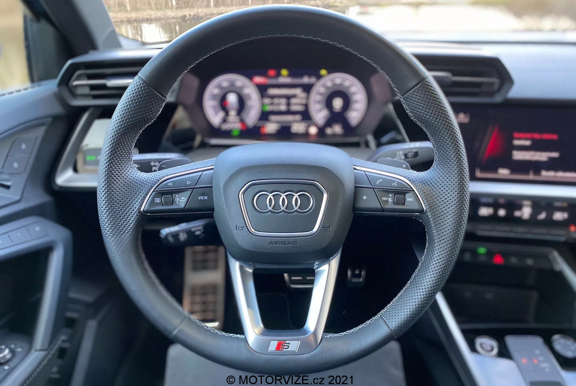 Pohľad z prvej osoby zo sedadla vodiča Audi A3 2020 (A3 Sportback), ktorý ukazuje športový trojramenný volant s logom Audi, multifunkčnými ovládacími tlačidlami, osvetlenou digitálnou prístrojovou doskou a časťou infotainment systému s médiami, navigáciou a nastaveniami vozidla. Vlastnosti zdôrazňujú interaktívnu technológiu a luxusné komponenty s dôrazom na zážitok z jazdy.