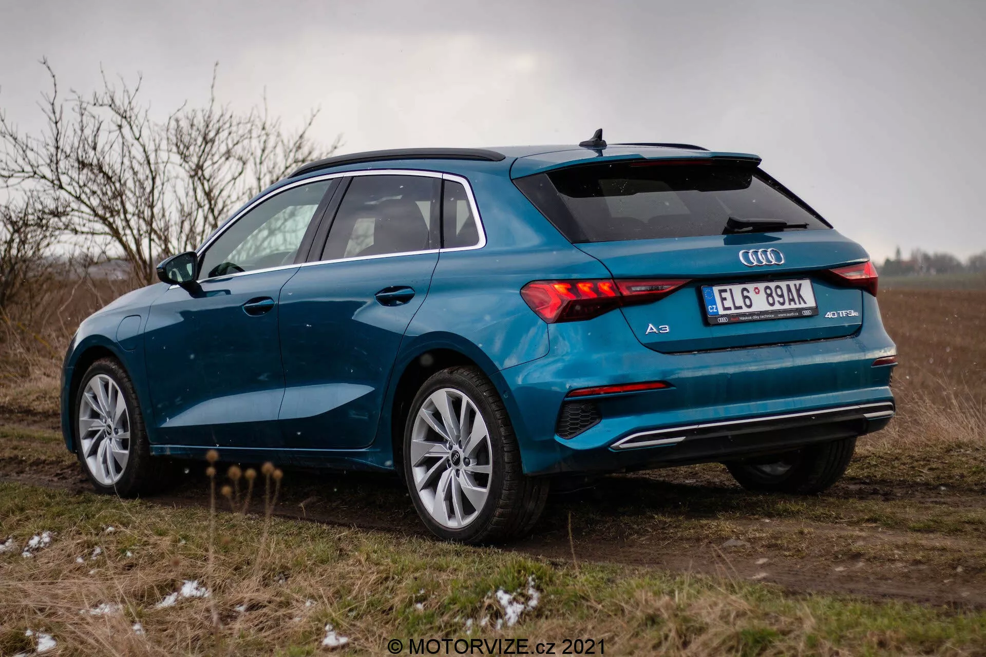 Modré Audi A3 2020 (Sportback) fotografované zo zadného trojštvrťového uhla na otvorenom poli pod zamračenou oblohou. Obrázok zvýrazňuje pravú stranu vozidla, zadné LED svetlá, dvojitý výfukový systém, viacramenné zliatinové kolesá a zadný spojler. Vozidlo je zaparkované na nespevnenej ploche s trsmi trávy a miernym množstvom snehu alebo námrazy, čo naznačuje chladné poveternostné podmienky. Celková nálada je ponurá s dôrazom na športový a prémiový dizajn Audi A3.