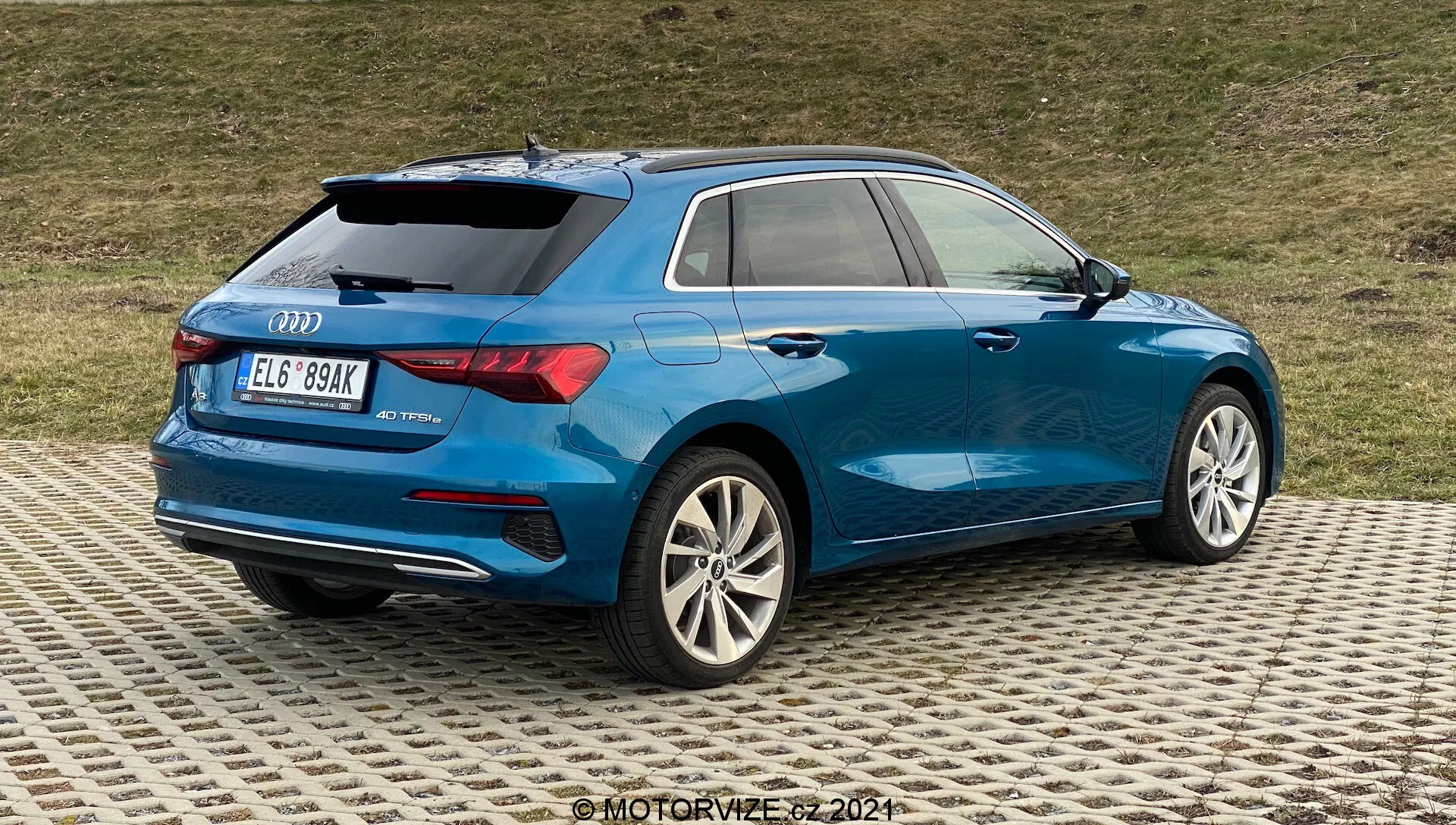Obrázok zobrazuje modrý Audi A3 2020 Sportback z trojštvrťového pohľadu zozadu, zdôrazňuje pravú zadnú stranu. Kľúčové detaily zahŕňajú viacramenné zliatinové kolesá so strieborným povrchom, čiastočne viditeľné zadné LED svetlá, elegantnú aerodynamickú karosériu s výraznou líniou ramien, zadné označenie naznačujúce model a možno aj úroveň vybavenia, tmavšie zadné okná pre súkromie, malý strešný spojler, strešné nosiče pre všestrannosť a dvojité výfukové rúry v spodnom zadnom nárazníku. Auto je zaparkované na vzorovanom povrchu s trávnatým pozadím, čo naznačuje, že prostredie nie je príliš mestské.
