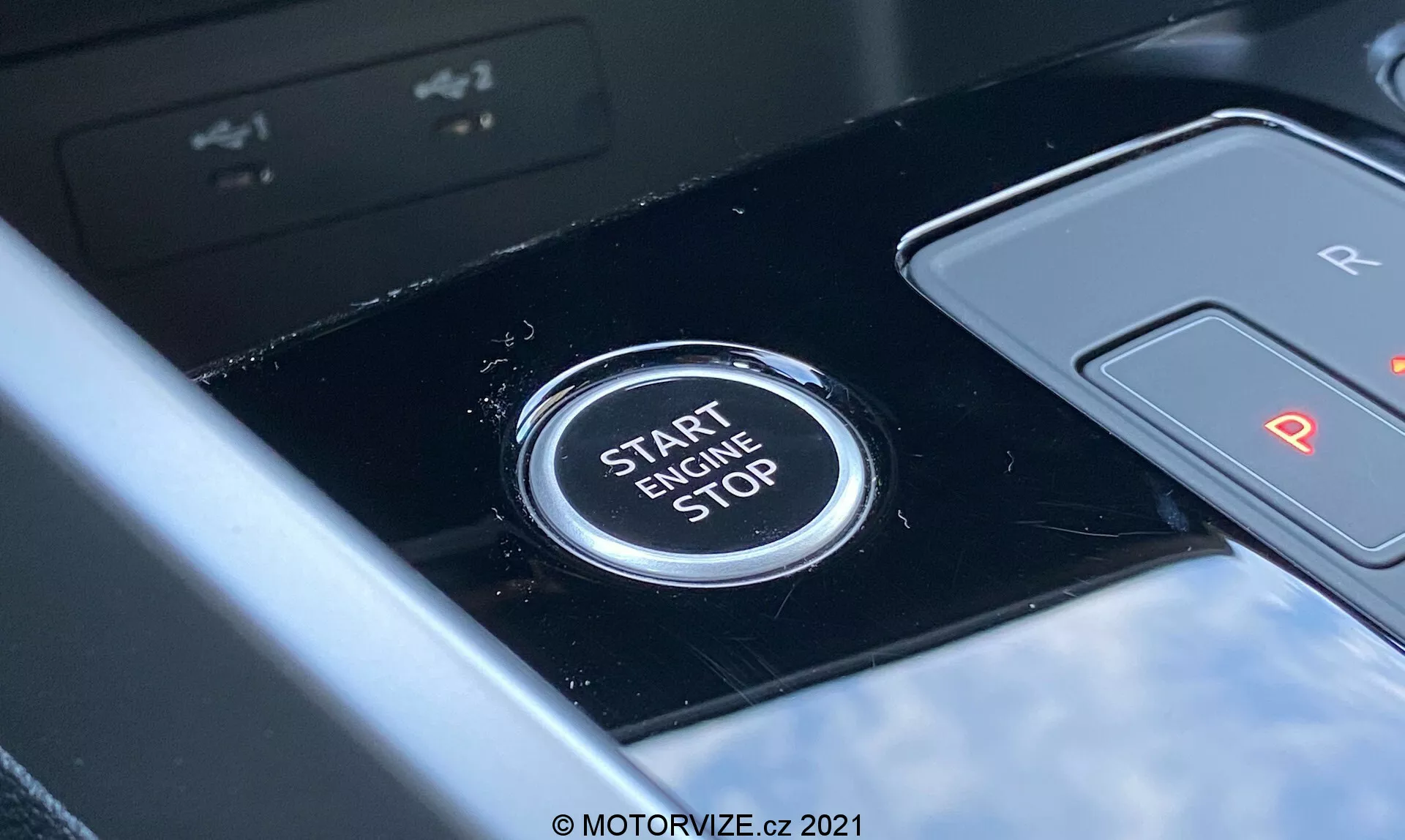 Detailný pohľad na tlačidlo štart/stop motora Audi A3 Sportback 2020 umiestnené v oblasti stredovej konzoly. Tlačidlo jasne zobrazuje text 'START ENGINE STOP' veľkými písmenami, čo naznačuje jeho funkciu na spustenie alebo vypnutie motora. Pohľadom z perspektívy vodiča, lesklý čierny panel a čiastočne viditeľná radiaca páka označená 'R' a 'P' naznačuje automatickú prevodovku. Plytká hĺbka ostrosti zdôrazňuje jasnosť tlačidla proti mierne rozmazanému pozadiu, čo poukazuje na moderný interiér vozidla s pokročilými technologickými funkciami.