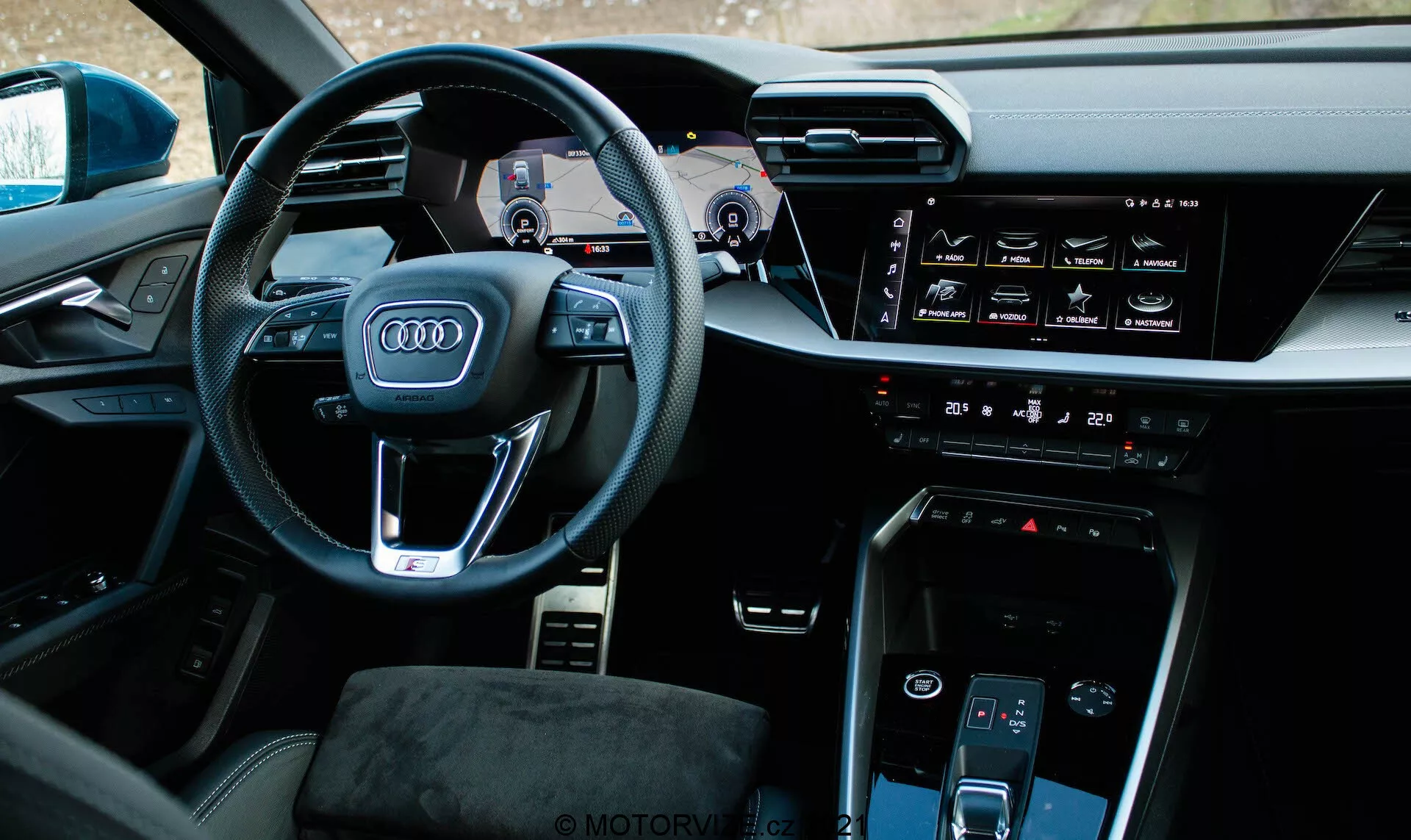 Pohľad do interiéru Audi A3 Sportback 2020 z mierne posunutého ľavého uhla zameraný na stranu vodiča. Výrazný volant nesie ikonické logo štyroch kruhov Audi, multifunkčné tlačidlá a viditeľné páčky radenia. Za volantom sa nachádza digitálny prístrojový panel nazývaný Audi Virtual Cockpit, ktorý zobrazuje tachometer, otáčkomer a navigačnú mapu. Stredová konzola obsahuje horizontálny dotykový infotainment displej, ovládanie klimatizácie, tlačidlo štart/stop motora, ďalší dotykový displej pre ovládanie klimatizácie a minimalistický volič prevodovky. Interiér sa vyznačuje kvalitnými materiálmi a technologicky integrovaným dizajnom s prémiovým dojmom.